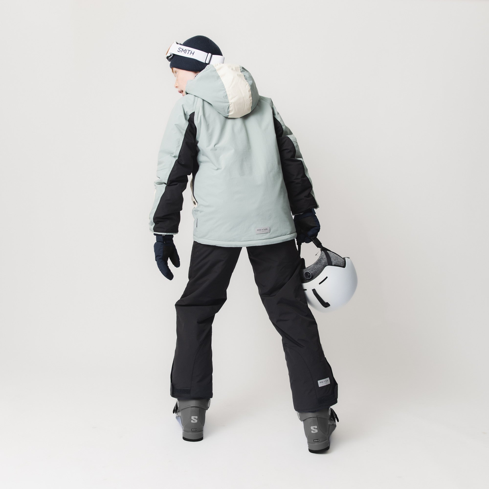 Child wearing MATWITTE snow pants in black paired with a pale blue ski jacket, holding a white helmet, viewed from behind to show the regular fit and how the trousers sit over ski boots during active use.