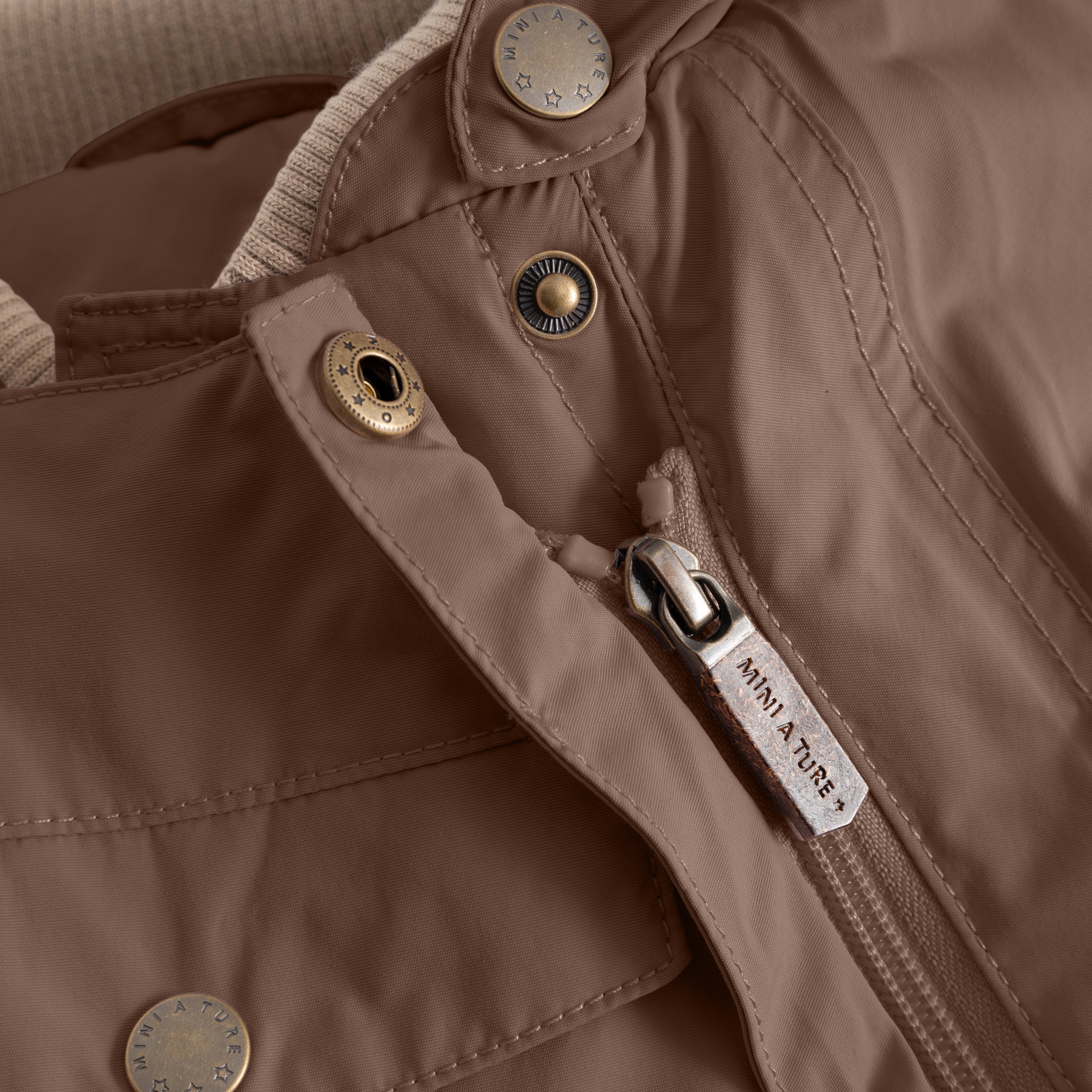 Close-up of the MATWISTI fleece lined snowsuit in warm brown showing the slanted front zip with branded MINI A TURE metal zip pull, embossed metal snap buttons and beige fleece lining visible at the collar opening.