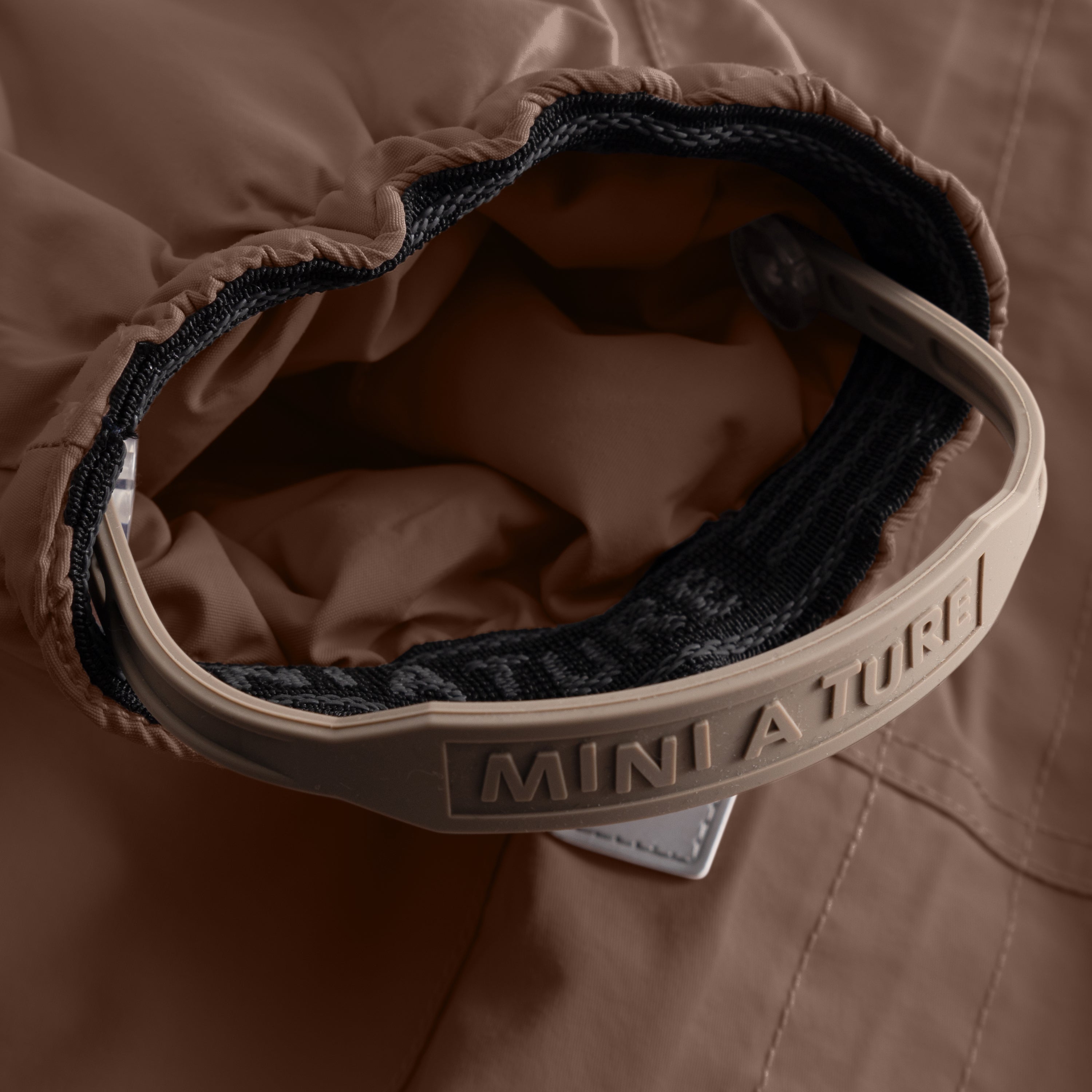 Close-up of the MATWISTI fleece lined snowsuit in warm brown showing the adjustable foot strap with embossed MINI A TURE branding on the beige plastic buckle, black elasticated cuff and soft fleece lining visible inside the leg opening.