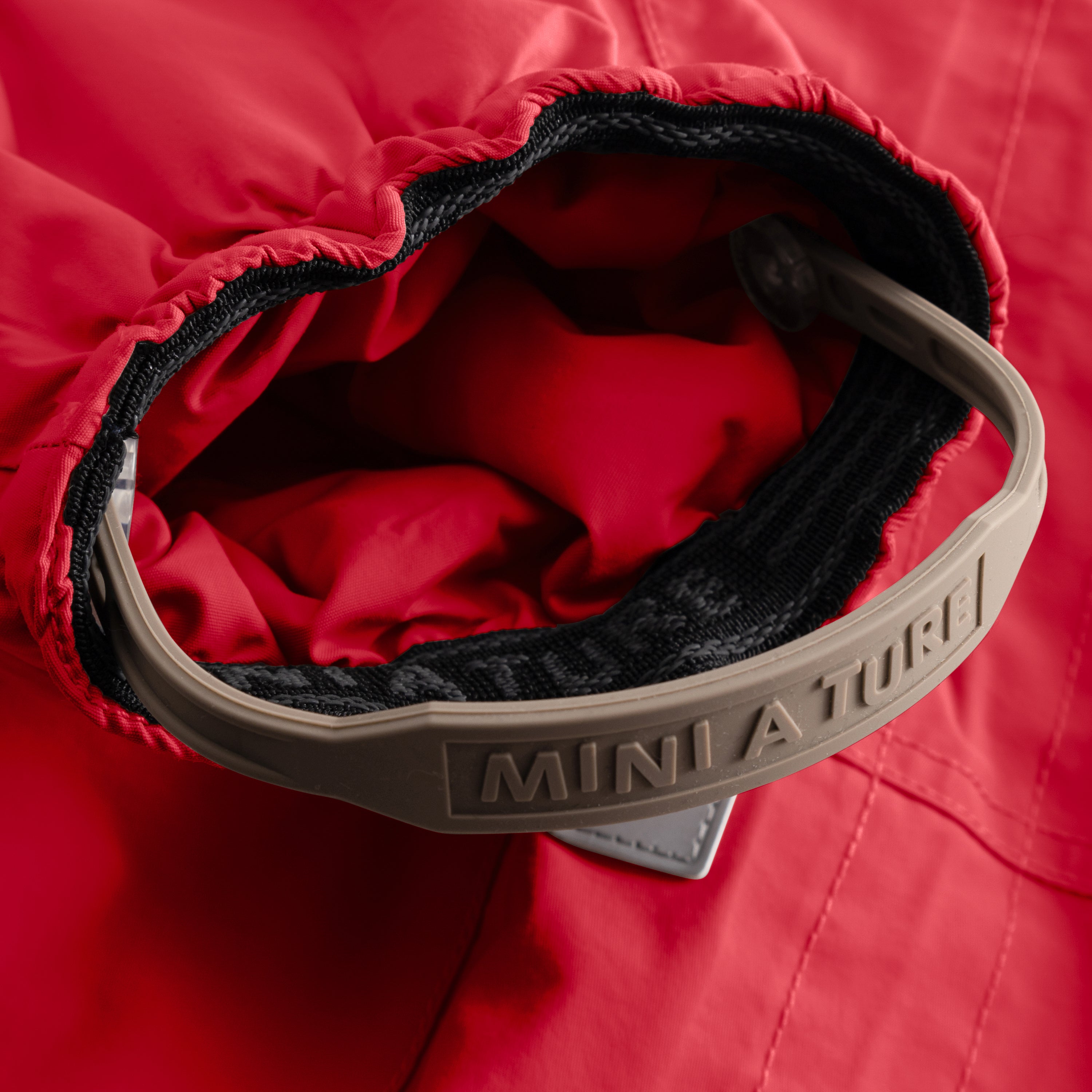 Close-up of the MATWISTI fleece lined snowsuit in bright red showing the adjustable foot strap with embossed MINI A TURE branding on the beige plastic buckle, black elasticated cuff and soft fleece lining visible inside the leg opening.