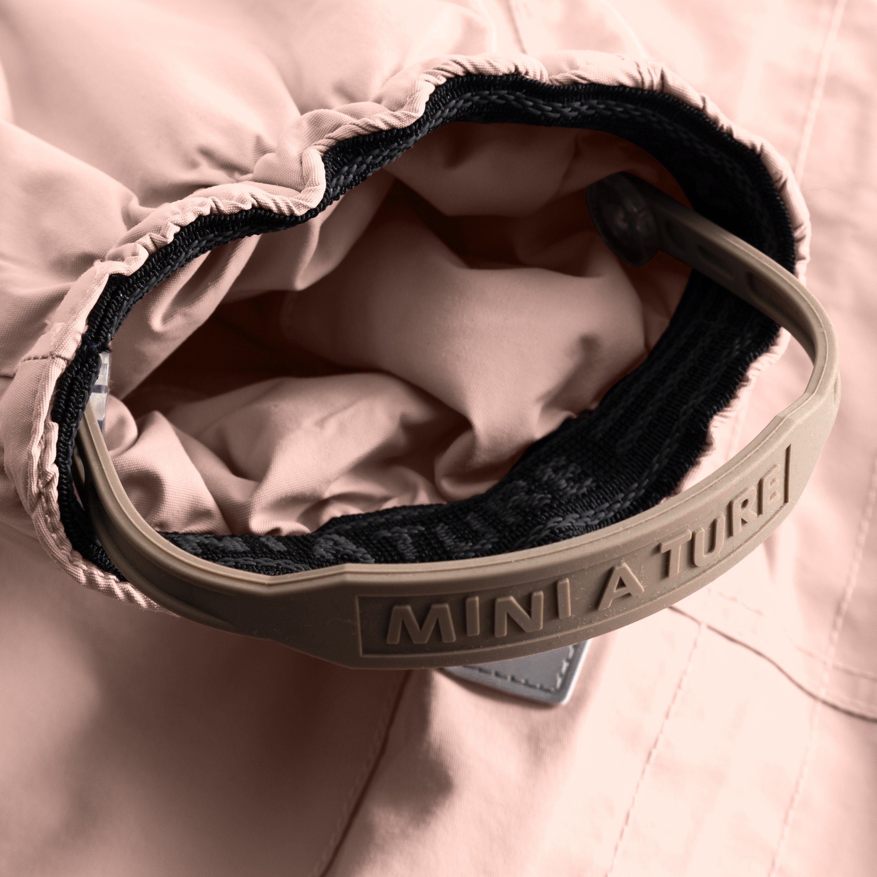 Close-up of the MATWISTI fleece lined snowsuit in soft rose pink showing the adjustable foot strap with embossed MINI A TURE branding on the beige plastic buckle, black elasticated cuff and soft fleece lining visible inside the leg opening.