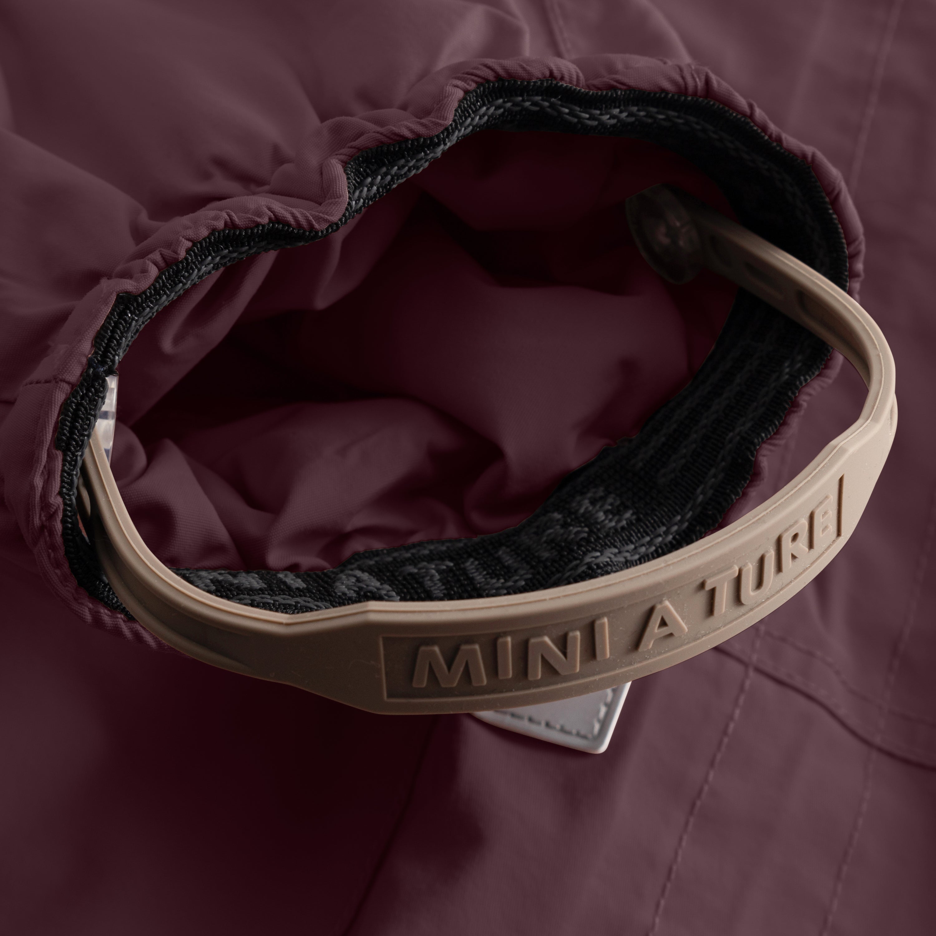 Close-up of the MATWISTI fleece lined snowsuit in deep plum showing the adjustable foot strap with embossed MINI A TURE branding on the beige plastic buckle, elasticated cuff and soft fleece lining visible inside the leg opening.