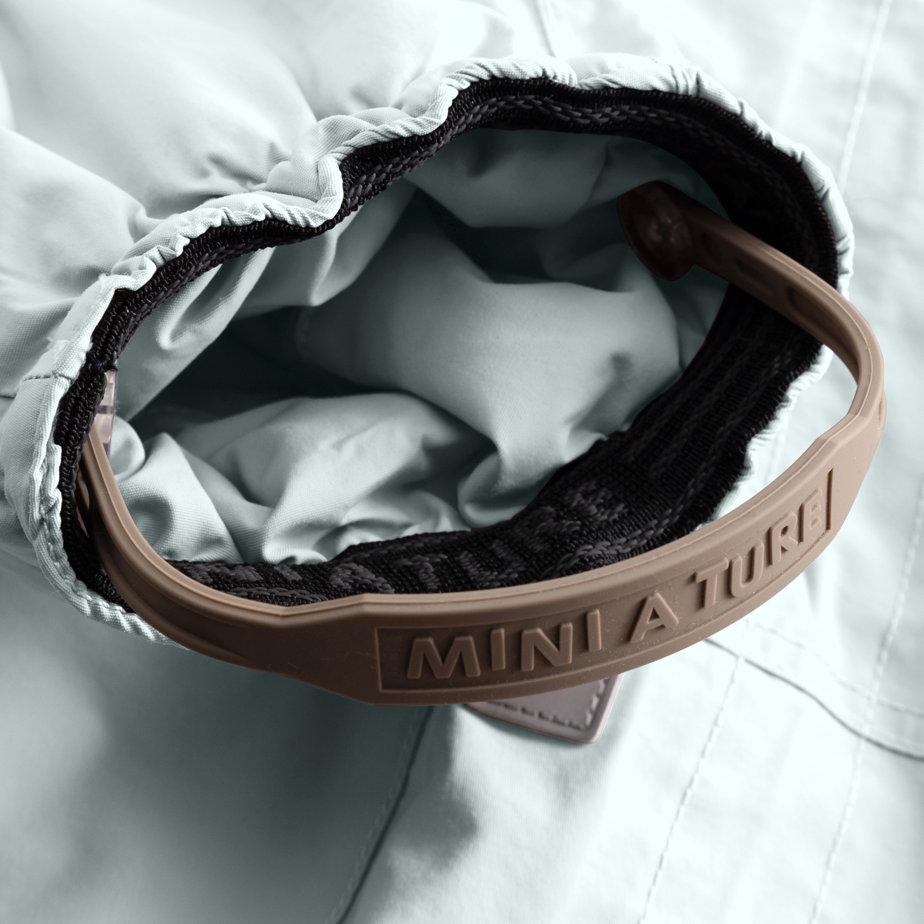 Close-up of the MATWISTI fleece lined snowsuit in pale blue showing the adjustable foot strap with embossed MINI A TURE branding on the beige plastic buckle, black elasticated cuff and soft fleece lining visible inside the leg opening.