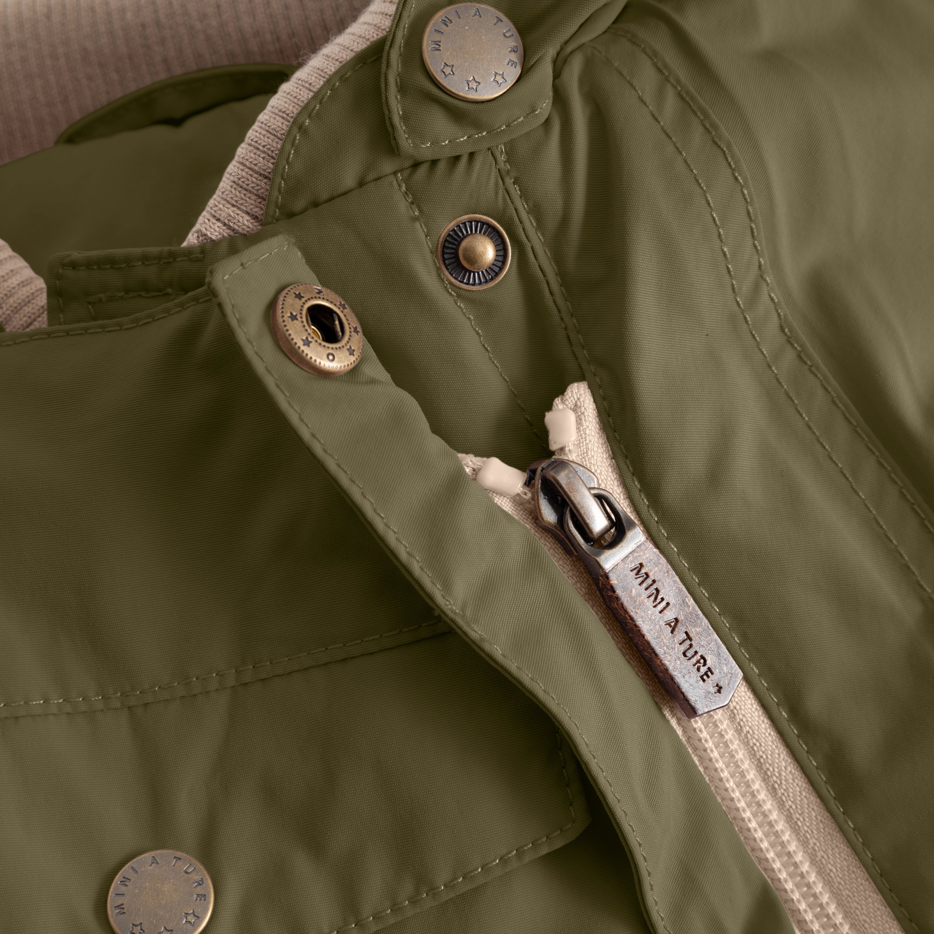 Close-up of the MATWISTI fleece lined snowsuit in olive green showing the slanted front zip with branded MINI A TURE metal zip pull, embossed metal snap buttons and beige fleece lining visible at the collar opening.
