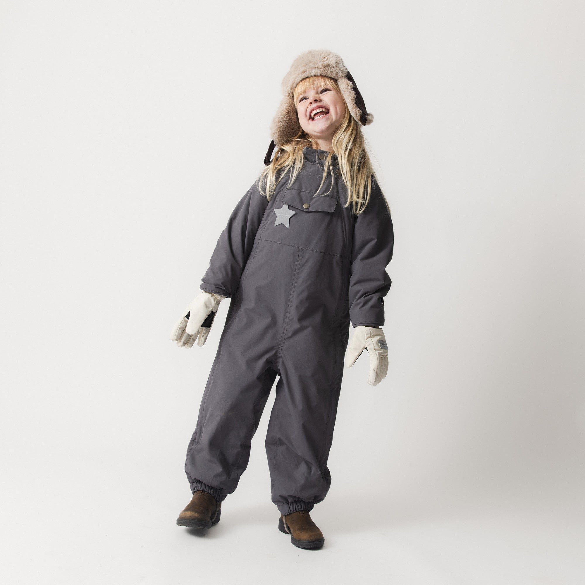 Young child wearing the MATWISTI fleece lined snowsuit in charcoal grey with a faux-fur trapper hat and cream mittens, showing the one-piece construction, front chest pocket with reflective star, elasticated cuffs and leg openings, and the suit's freedom of movement during active play.