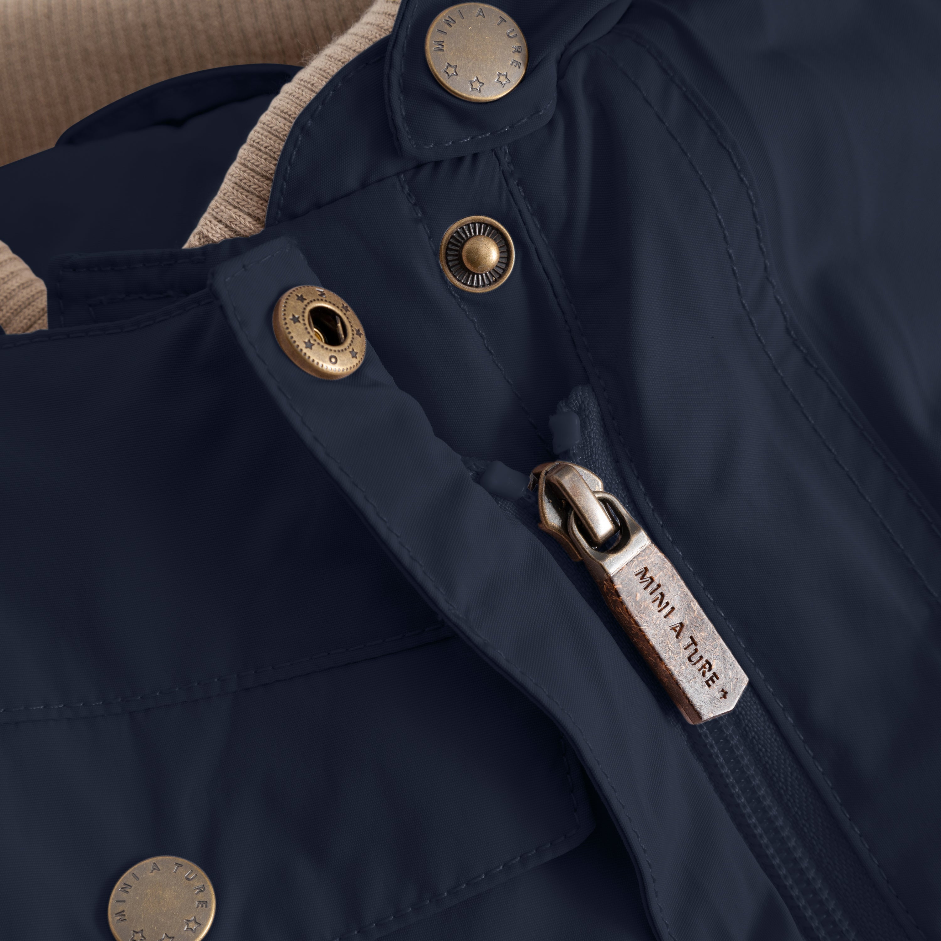 Close-up of the MATWISTI fleece lined snowsuit in navy blue showing the slanted front zip with branded MINI A TURE metal zip pull, embossed metal snap buttons and beige fleece lining visible at the collar opening.