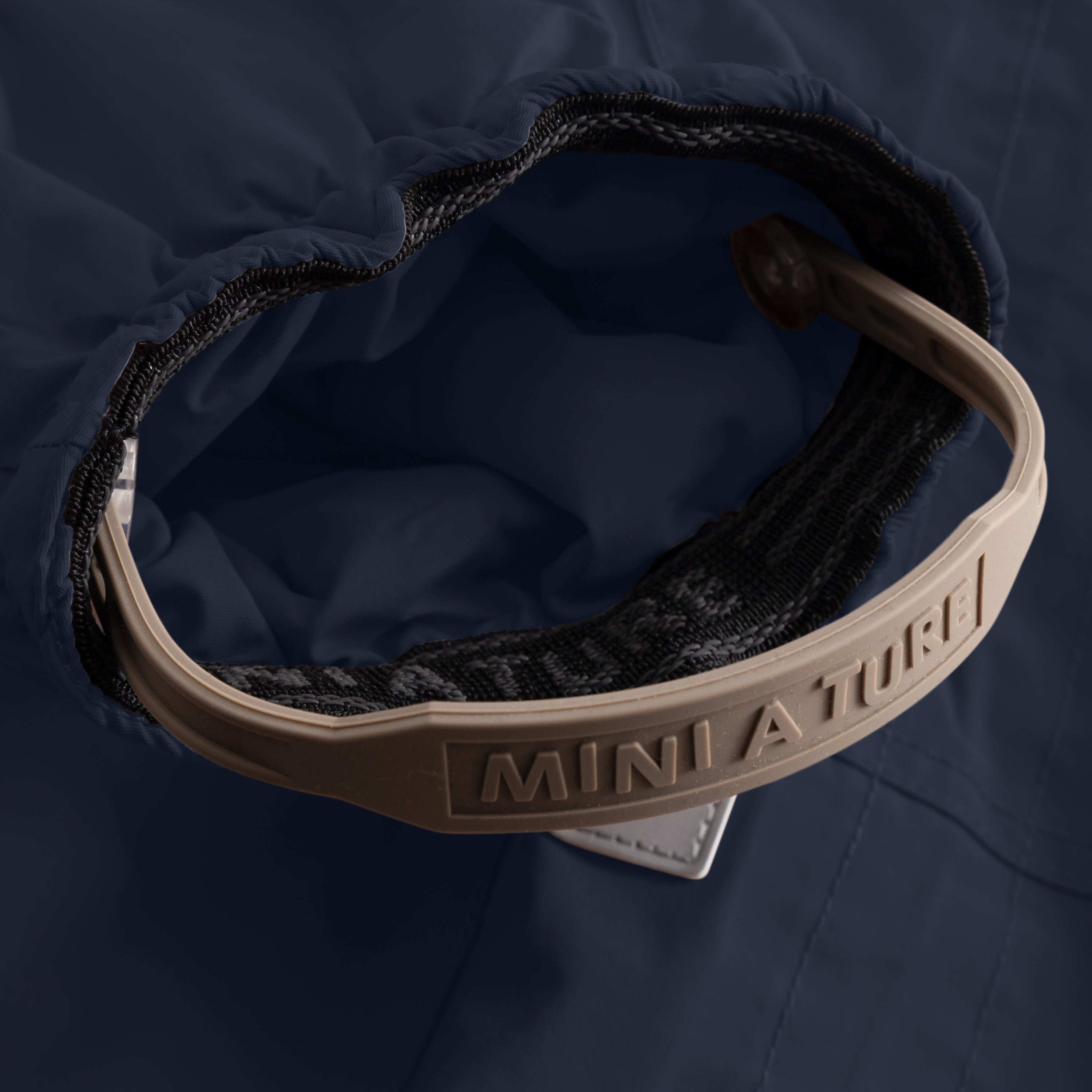 Close-up of the MATWISTI fleece lined snowsuit in navy blue showing the adjustable foot strap with embossed MINI A TURE branding on the beige plastic buckle, black elasticated cuff and soft fleece lining visible inside the leg opening.