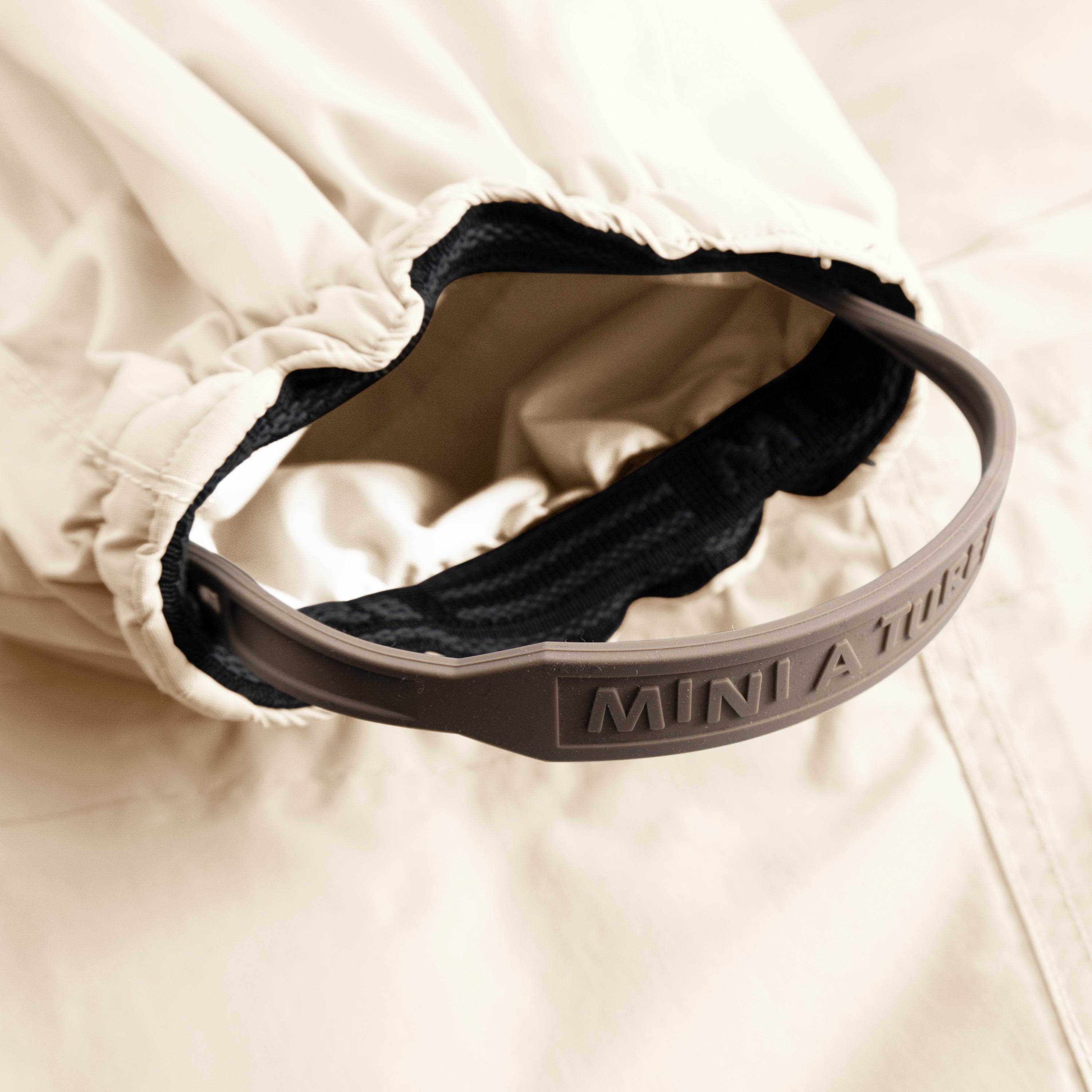 Detail of MATWISTI fleece lined snowsuit fur in cream showing the adjustable and detachable foot strap with embossed MINI A TURE branding on the taupe silicone band, elasticated ankle cuff with beige fleece lining, and dark fleece interior visible against the waterproof outer shell.