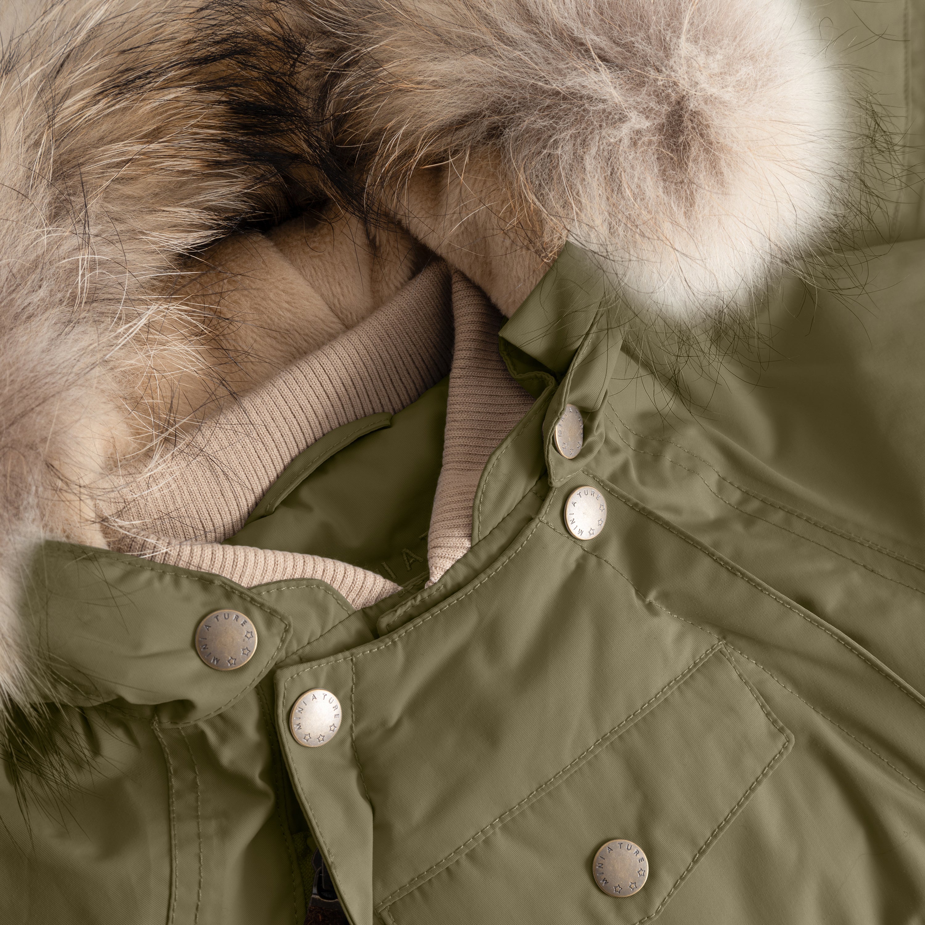 Close-up of MATWISTI fleece lined snowsuit fur in olive green showing the detachable genuine fur hood trim, ribbed taupe collar, MINI A TURE branded press-stud fastenings along the chest, and front pocket detail on the waterproof shell.