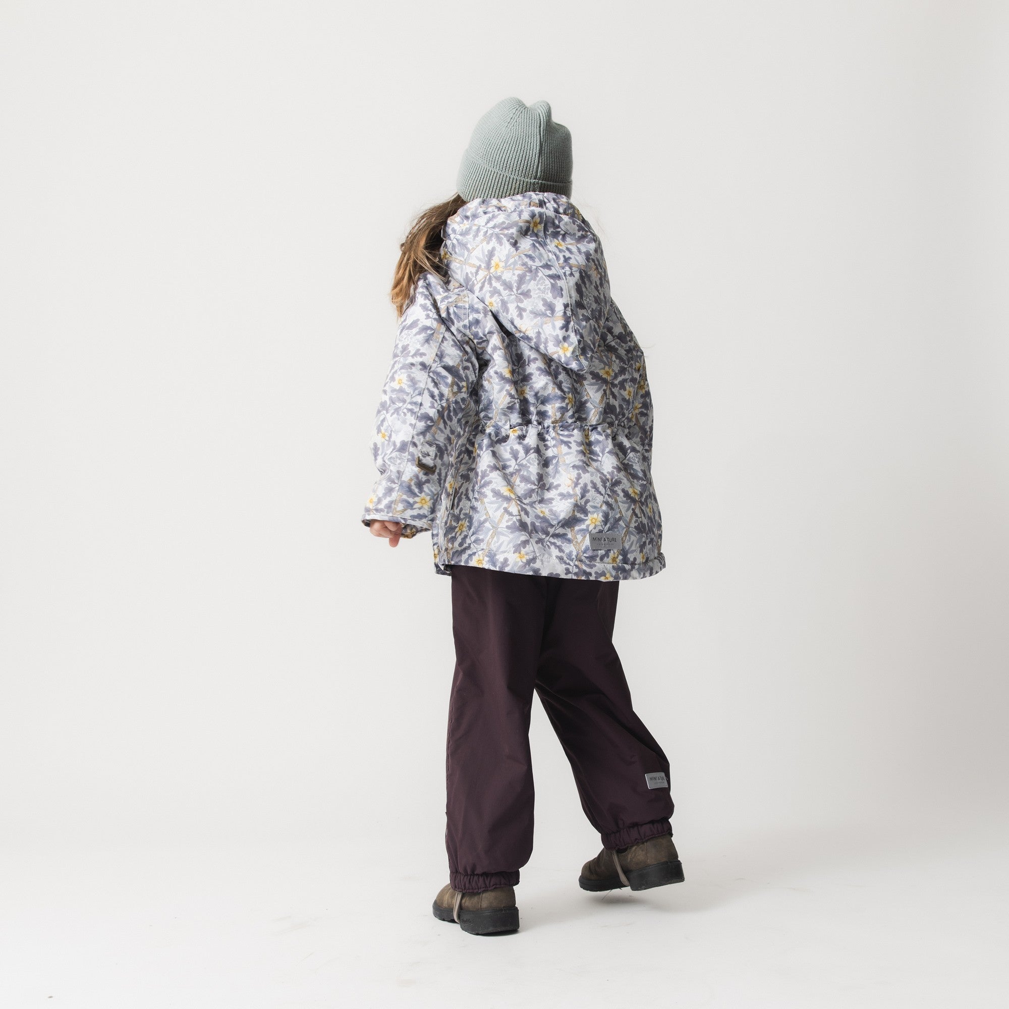 MATWANG fleece lined winter jacket seen from behind on a child wearing a grey knitted beanie and dark trousers, showing the full Scandinavian floral print, the adjustable fit through the torso, and the jacket's length paired with waterproof winter trousers.