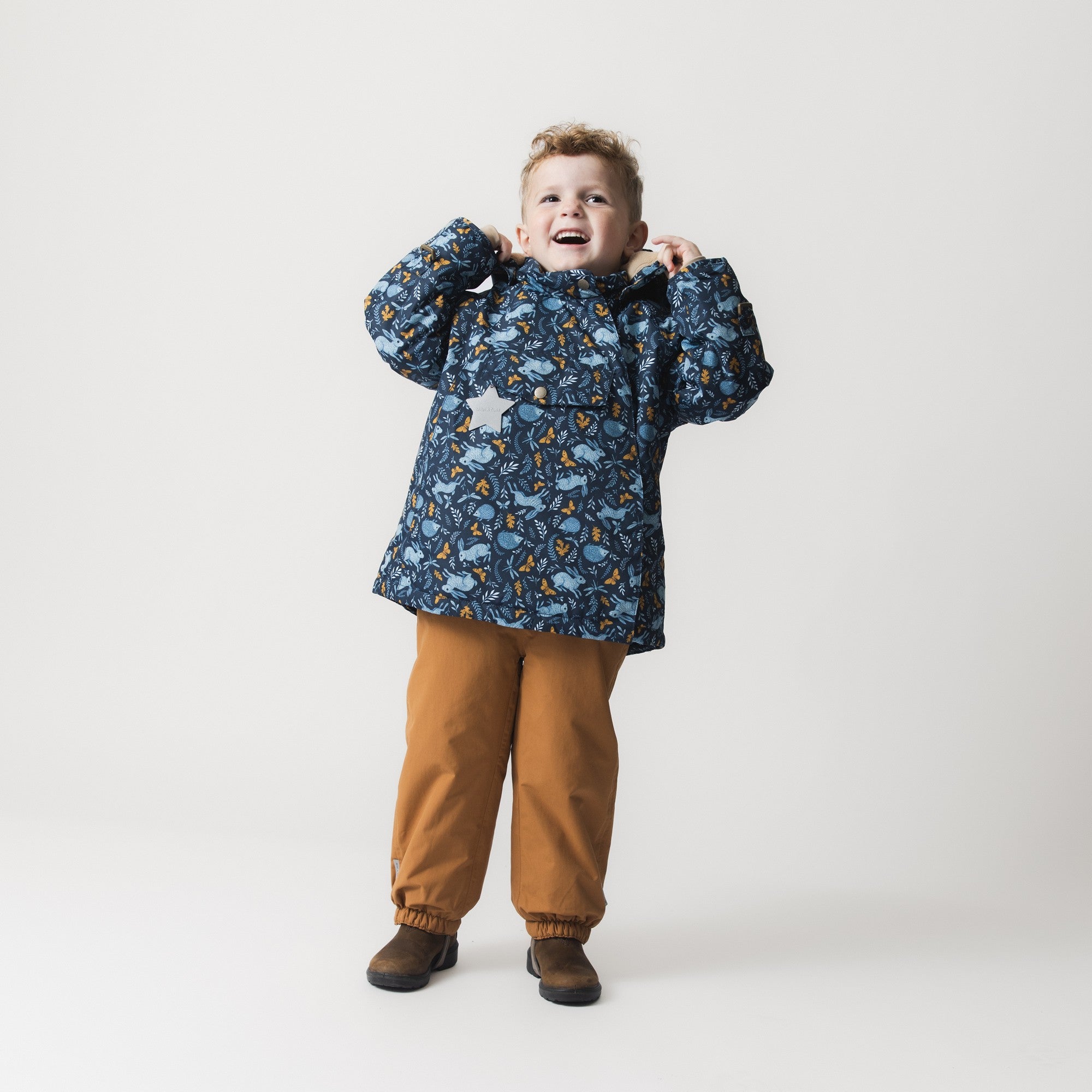 MATWANG fleece lined winter jacket worn by a young child in a joyful pose with hands raised to the hood, paired with mustard trousers, illustrating the jacket's relaxed fit and the Scandinavian floral print featuring blue rabbits and golden oak leaves across the waterproof shell.
