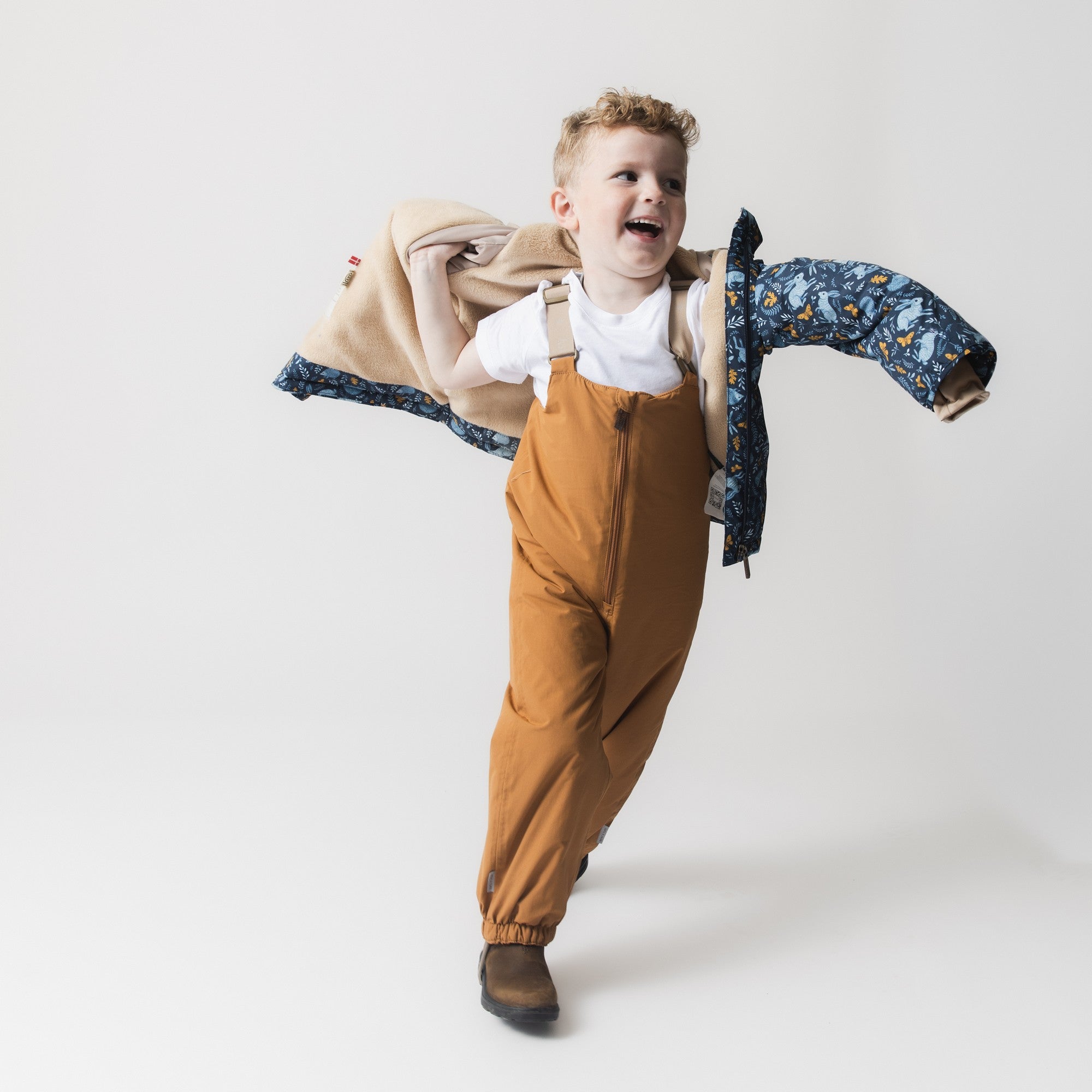 MATWANG fleece lined winter jacket held aloft by a child in mustard dungarees, capturing the jacket mid-movement to reveal both the Scandinavian floral print shell and the beige fleece lining visible inside the hood and body, demonstrating the jacket's lightweight construction and ease of handling.