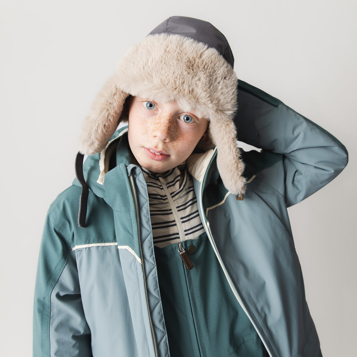 MATADANE ski tech jacket worn by a child adjusting a faux-fur trapper hat, shown in teal colourblock design with hood, front zip, reflective trim detail and ripstop recycled nylon shell over a striped base layer.