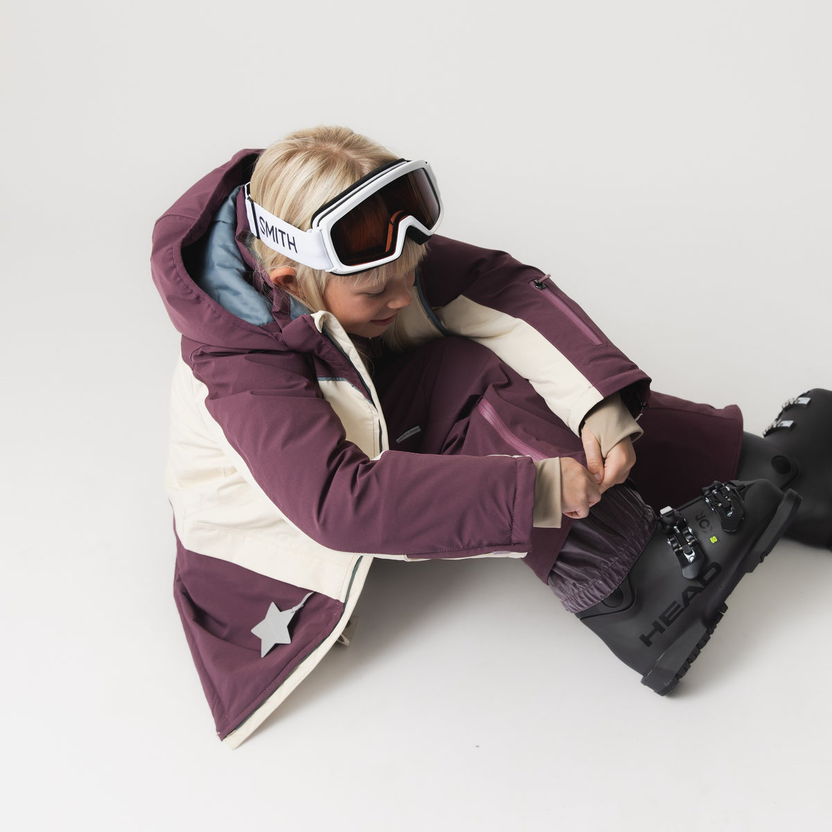 Kind in auberginefarbener MATADAL-Skihose mit beigen Einsätzen und weißer Skibrille sitzt und schließt den seitlichen Reißverschluss am Beinende über dem Skischuh.