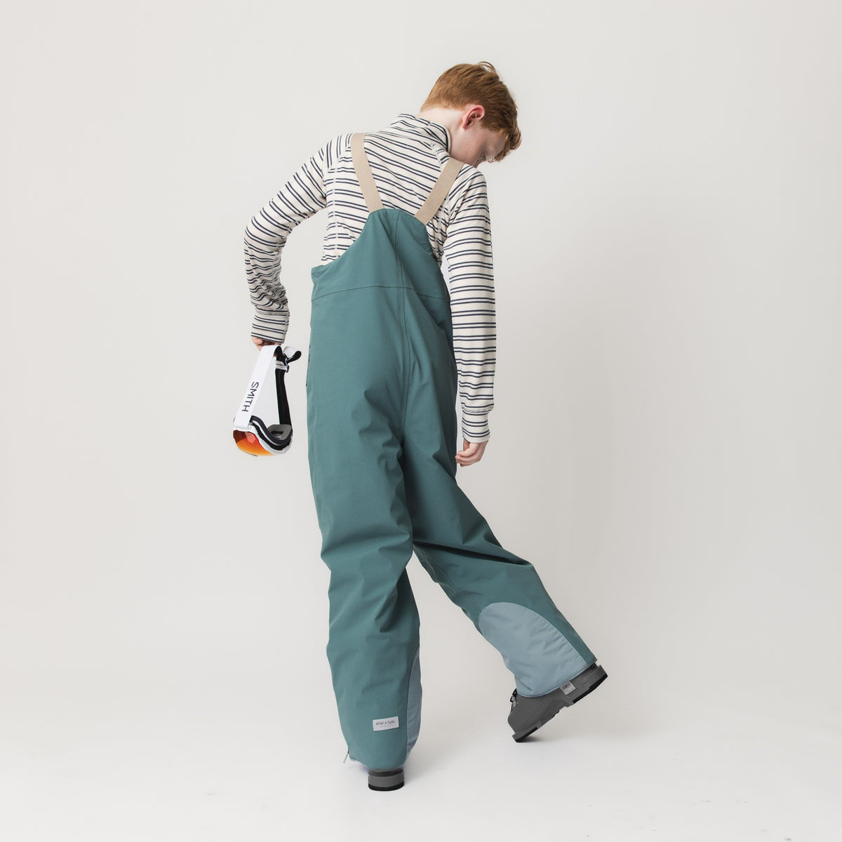 Kind in blaugrüner MATADAL-Skihose mit beigen Trägern und gestreiftem Langarmshirt hält ein Spielzeug und zeigt die wasserdichte Ripstop-Konstruktion mit Verstärkungen am unteren Hosenbein.