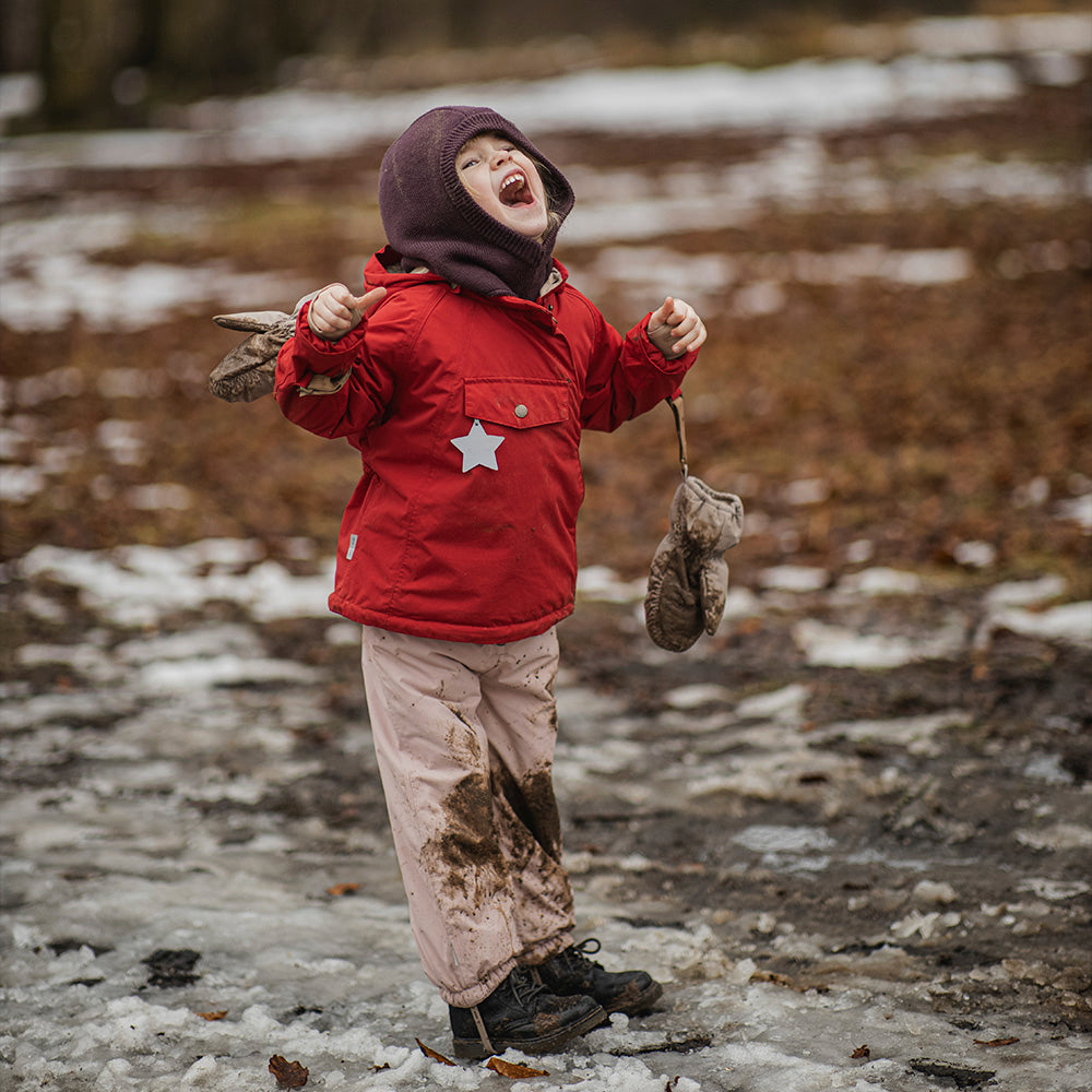 Child laughing joyfully outdoors in the MATWANG fleece-lined winter jacket in red with detachable pointed hood, paired with a dark knitted beanie and pale pink trousers, holding mittens whilst standing in muddy, snowy terrain.