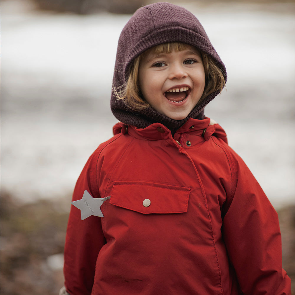 Young child smiling outdoors in the MATWANG fleece-lined winter jacket in red with a detachable pointed hood worn up, paired with a purple knitted beanie, showing the waterproof recycled nylon shell and reflective star detail on the chest pocket.