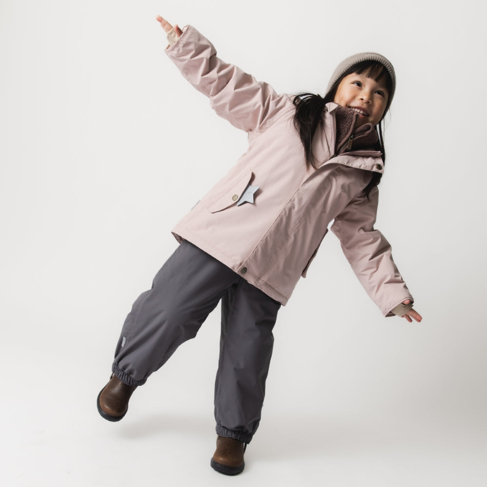 Young child wearing the MATWALLY fleece lined winter jacket in dusty pink paired with charcoal grey trousers and a beige knitted hat, captured mid-movement with one arm raised and a joyful expression, demonstrating the jacket's freedom of movement and regular fit.