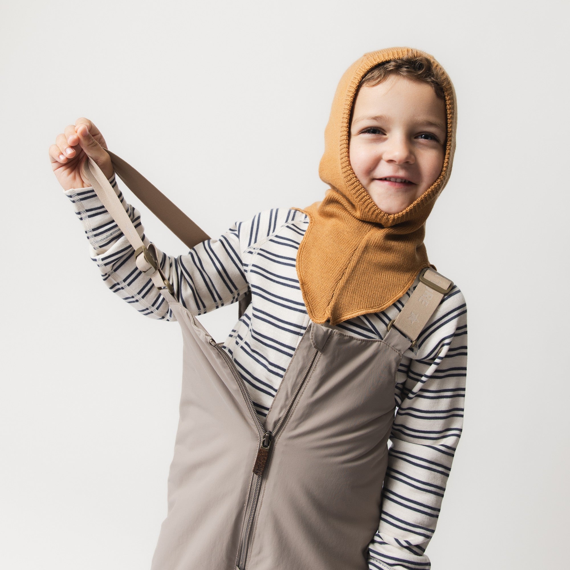 MATKEA light balaclava in golden ochre merino wool, worn by a smiling child over a striped top and dungarees, illustrating the hood's close fit around the face and the extended shoulder coverage that layers neatly beneath everyday clothing.
