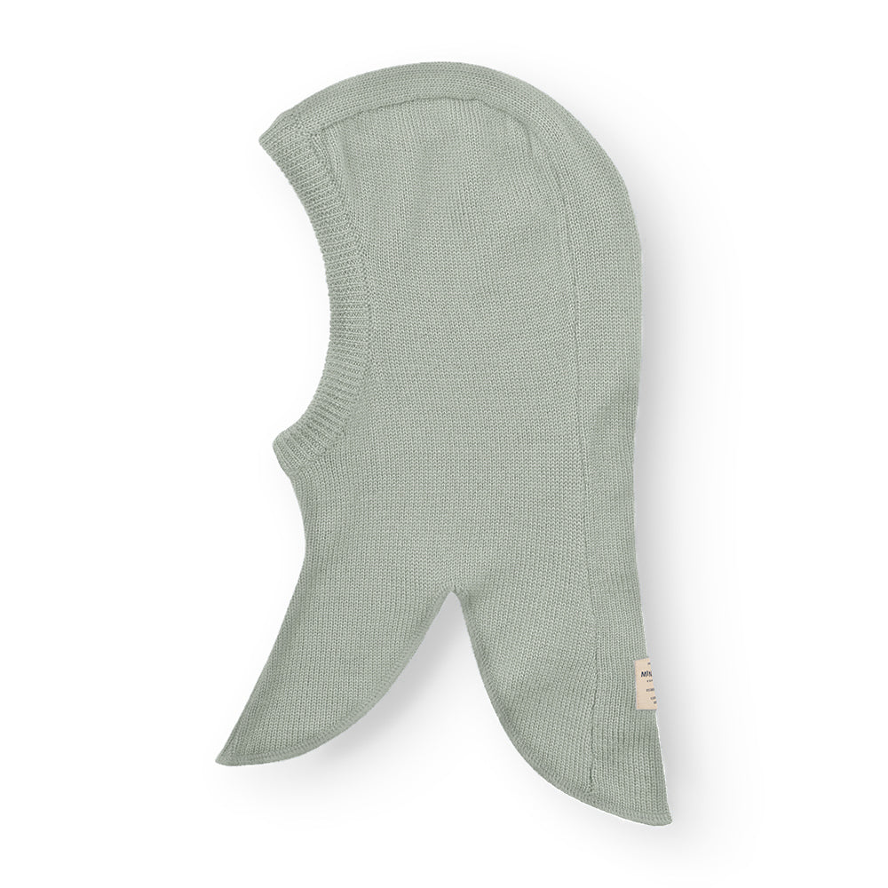 MATKEA light balaclava in sage green merino wool, photographed flat to reveal the smooth knitted texture and distinctive pointed shoulder flaps that extend protection down the upper back and chest.