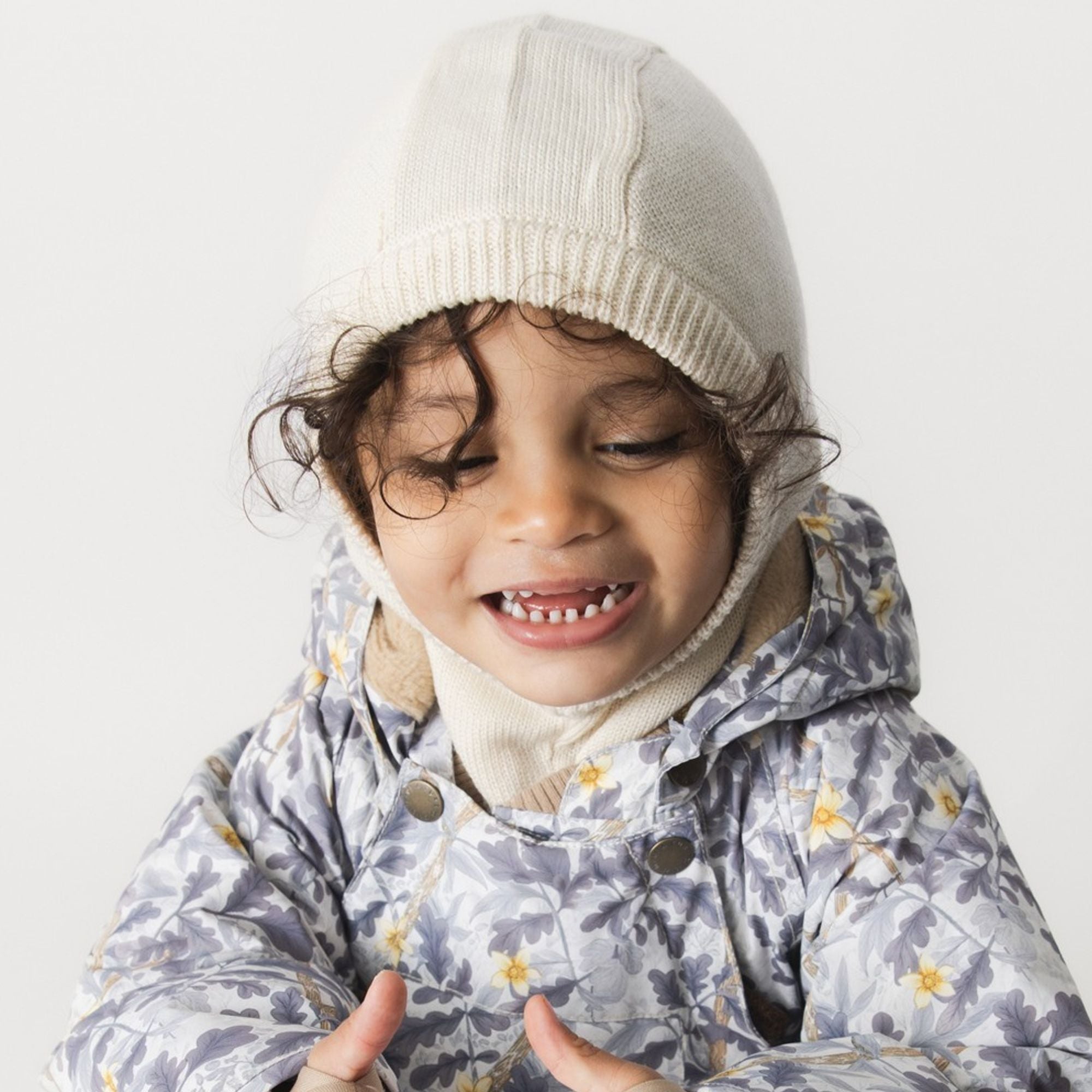 MATKEA light balaclava in cream merino wool, worn by a smiling toddler over a floral jacket, demonstrating the snug fit around the face and neck with the ribbed edge sitting comfortably at the forehead.