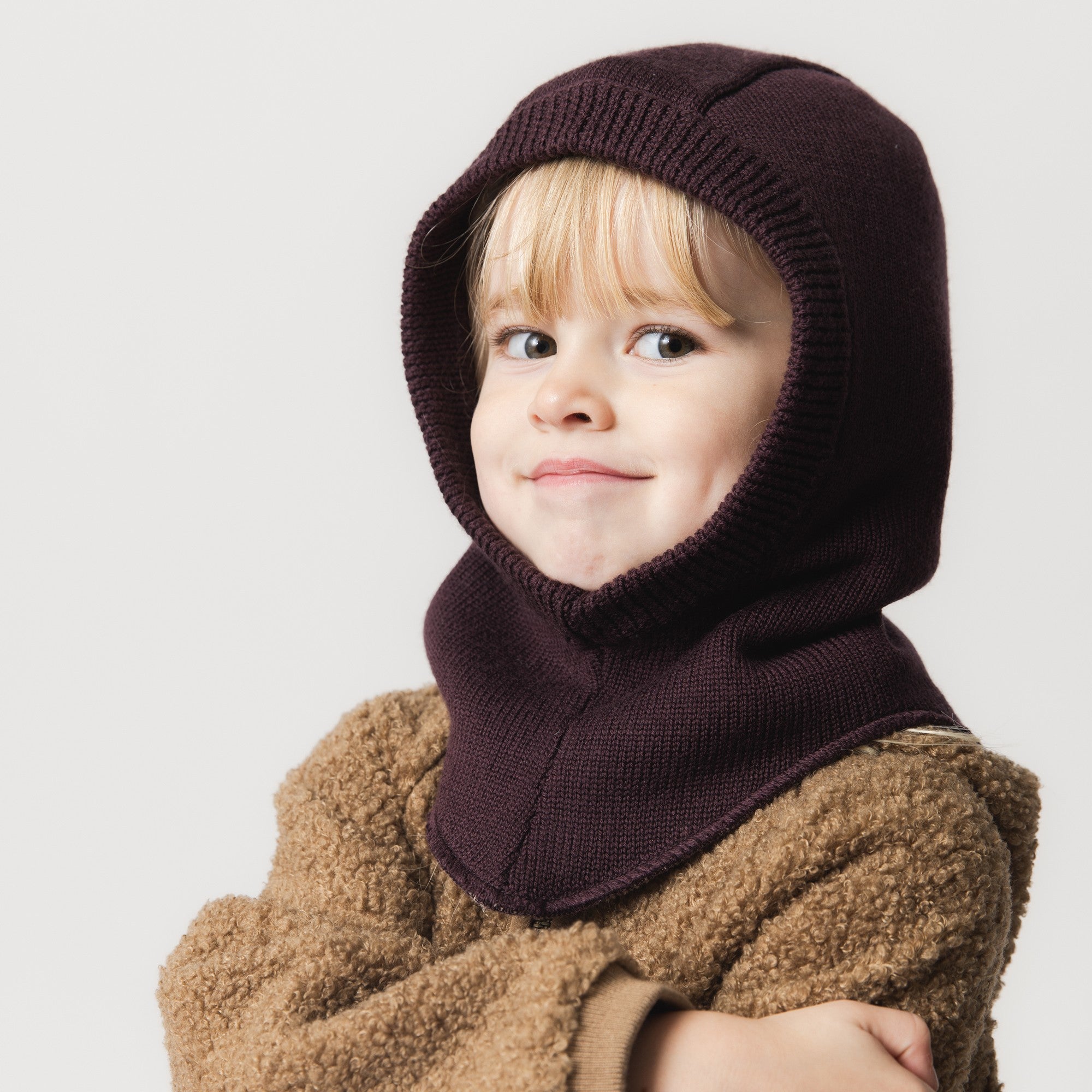 Child wearing the MATJORK balaclava in deep plum, showing how the merino wool and fleece lining wrap the head and neck in one layer, with the face opening framed by soft ribbed knit.