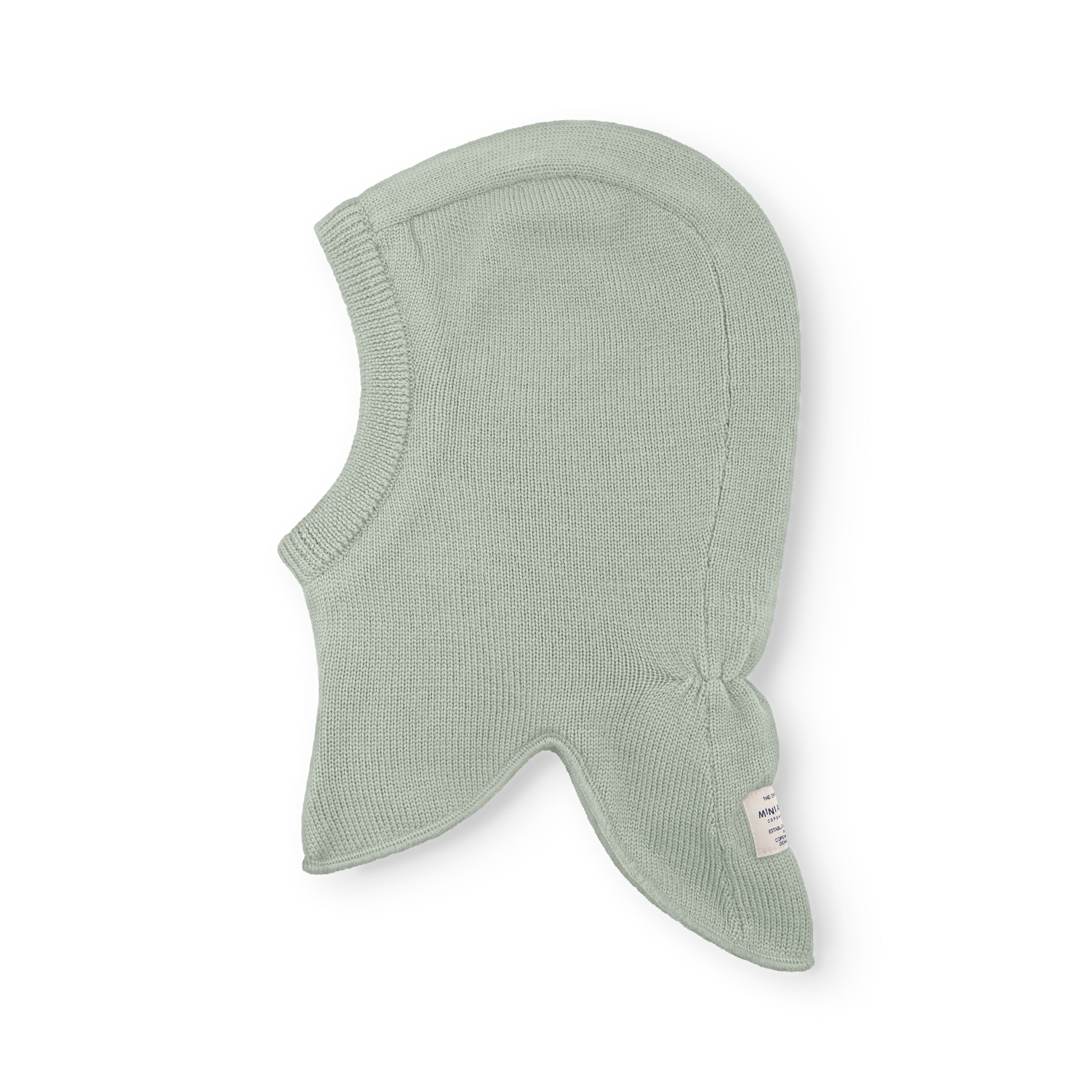 MATJORK balaclava in pale sage green merino wool shown flat, displaying the ribbed knit texture, face opening and extended neck coverage, with the MINI A TURE label visible at the lower right.