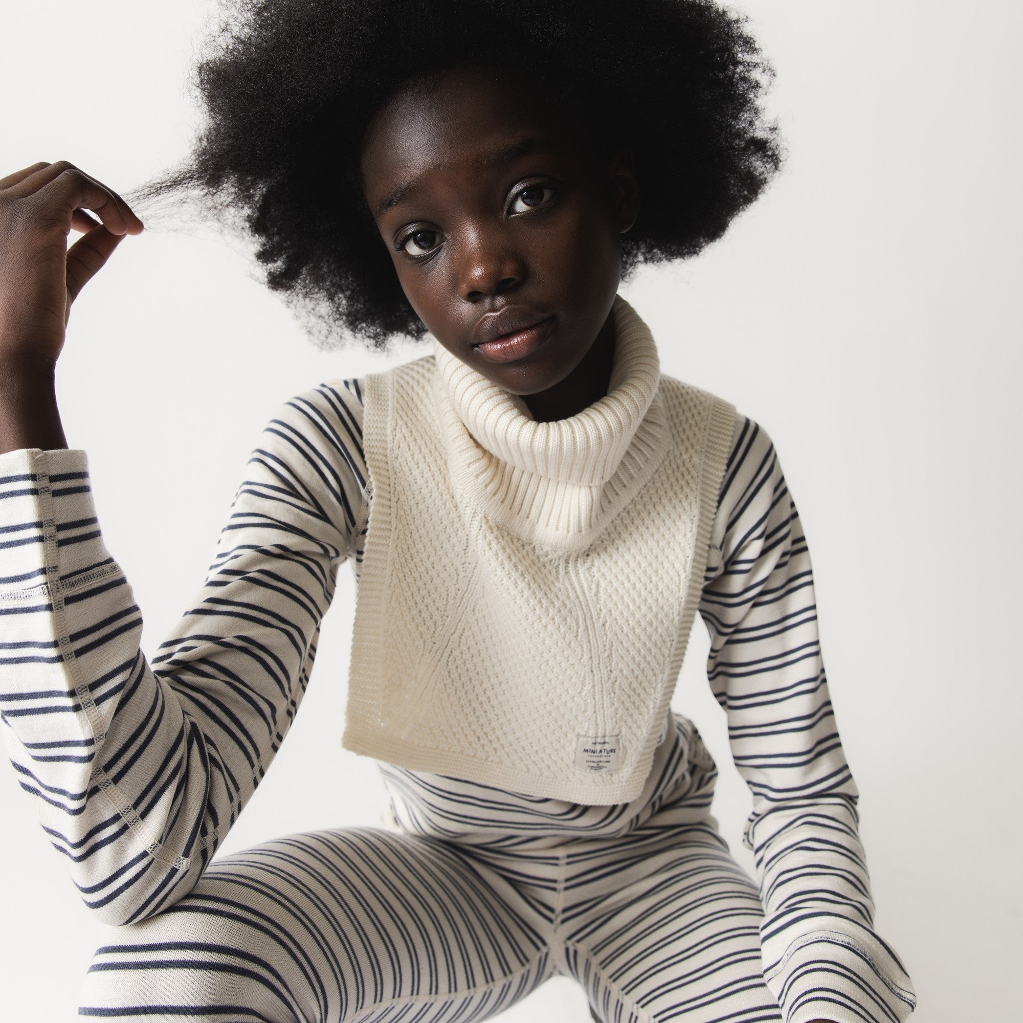 MATTAYLER neckwarmer in cream merino wool, worn by a child over a navy and cream striped long-sleeve top, showing the wide ribbed high neck and textured front panel sitting close to the throat and upper chest.
