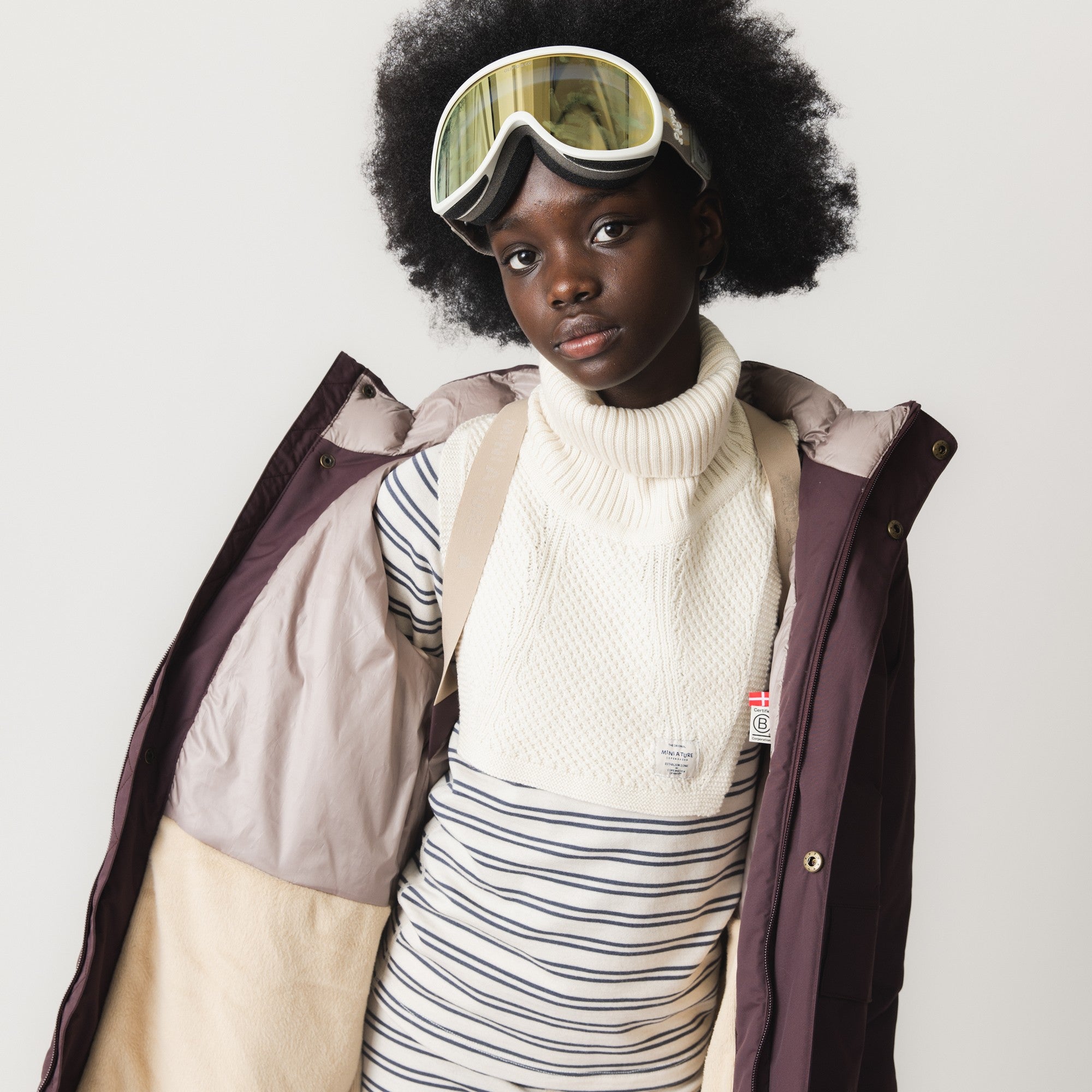 MATTAYLER neckwarmer in cream merino wool, worn by a child with ski goggles on the forehead and a purple and beige ski jacket open to reveal the wide ribbed high neck and textured front panel layered over a navy and cream striped base layer.