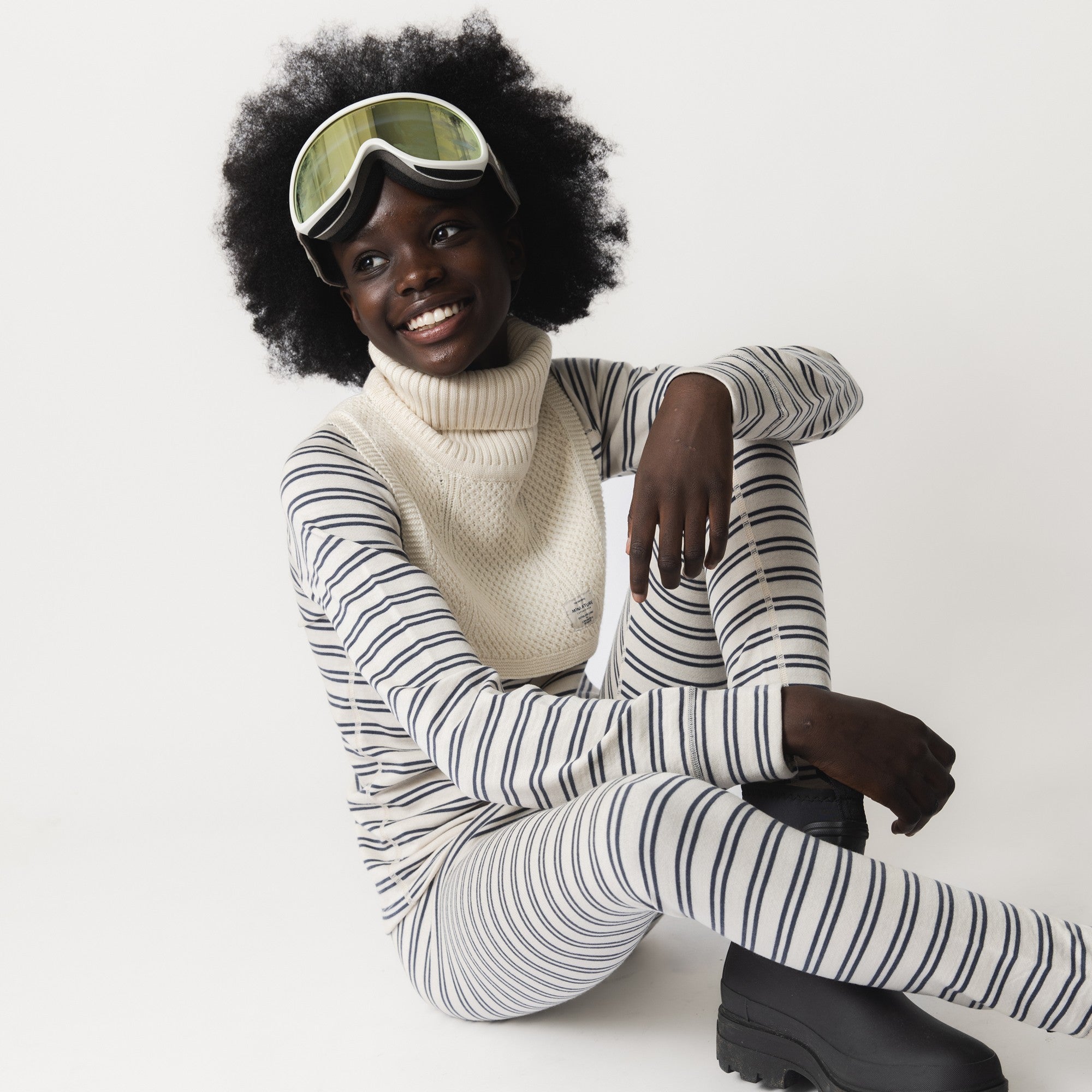 MATTAYLER neckwarmer in cream merino wool, worn by a child seated on the floor with ski goggles pushed up onto the forehead, layered over a navy and cream striped thermal base layer, showing the wide ribbed high neck and textured front panel.