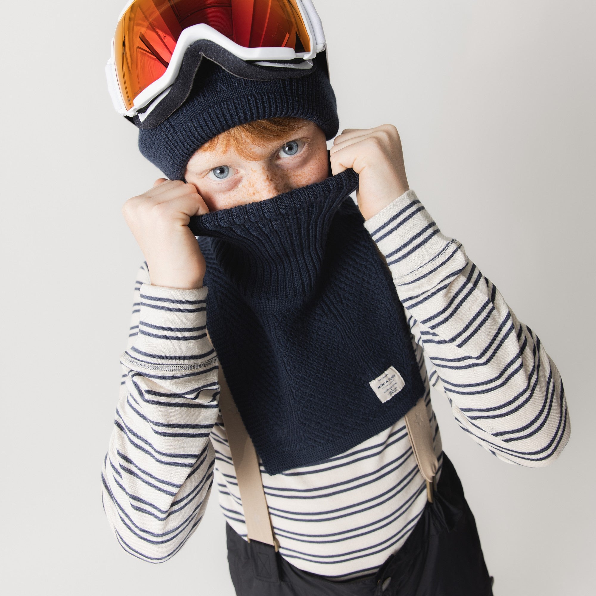MATTAYLER neckwarmer in navy blue merino wool, shown pulled up over a child's nose and mouth, with a high neck and extended front panel providing full coverage beneath a striped top and ski goggles.
