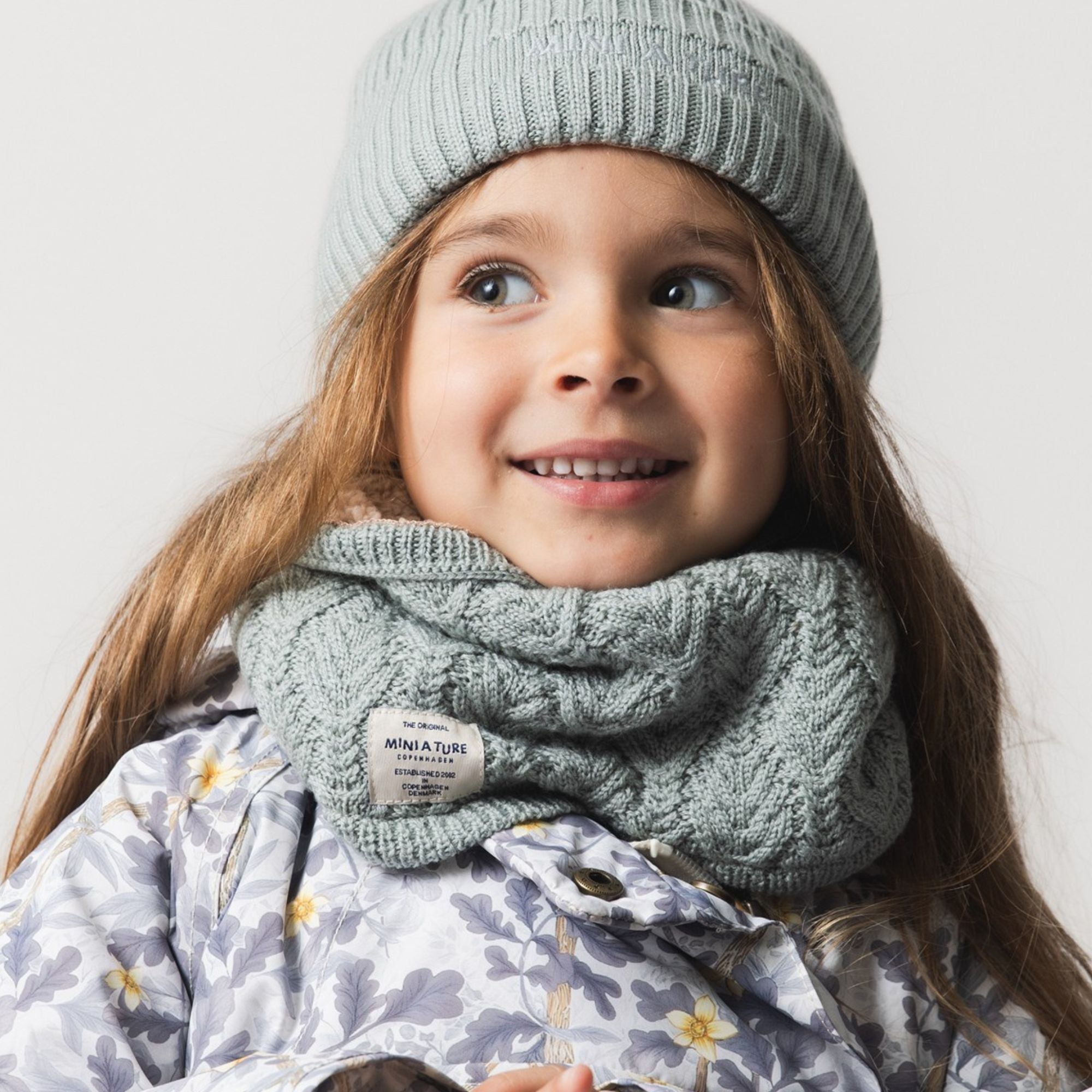 MATGOI fleece lined neckwarmer in soft grey merino wool with structured knit pattern, worn by a smiling child with a matching beanie and floral jacket.