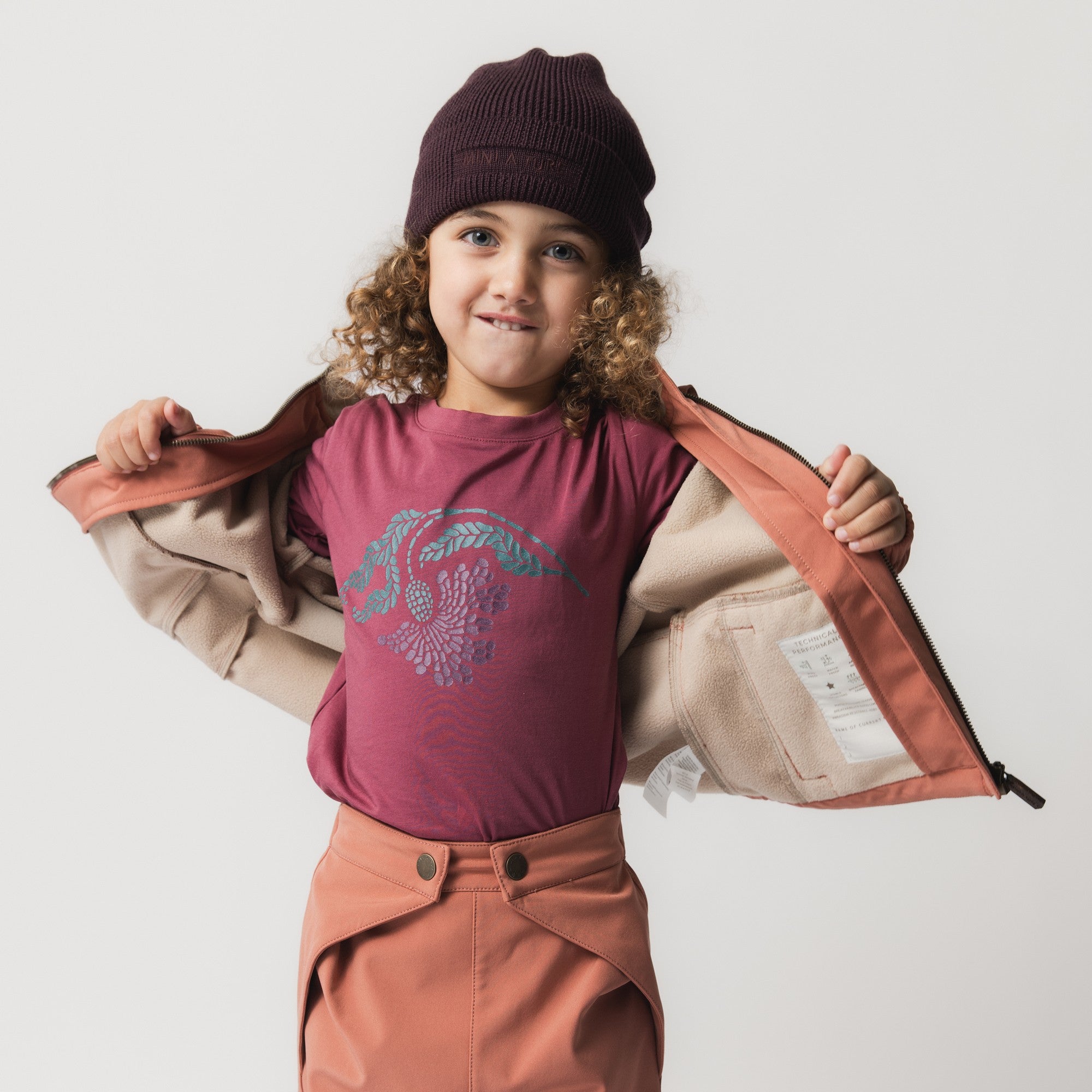 MATBOJE beanie in deep plum merino wool with a ribbed knit and slouched fit, worn by a smiling child in a burgundy t-shirt and coral fleece-lined jacket held open to show the interior.
