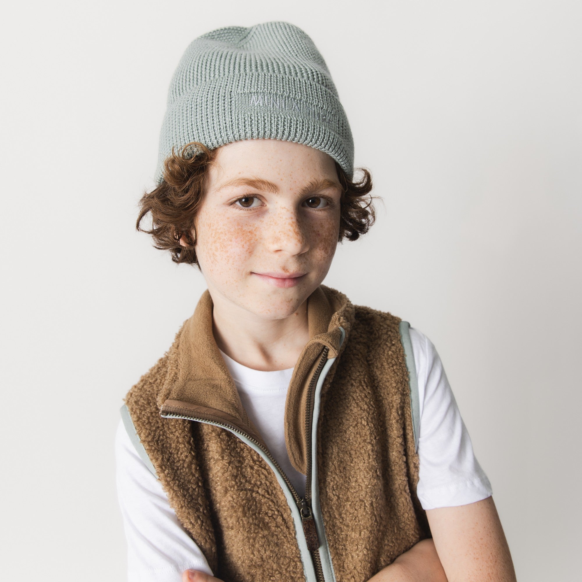 MATBOJE beanie in soft grey-green merino wool with a ribbed knit and fold-up brim, worn by a child in a brown fleece gilet and white t-shirt.