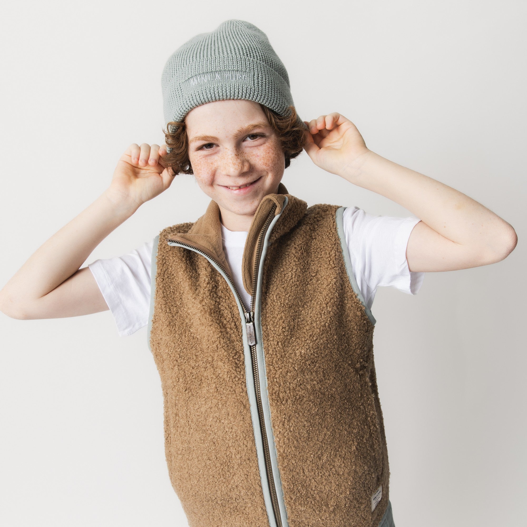 MATBOJE beanie in pale sage merino wool with a ribbed knit, worn by a smiling child in a fleece gilet who is adjusting the hat with both hands.