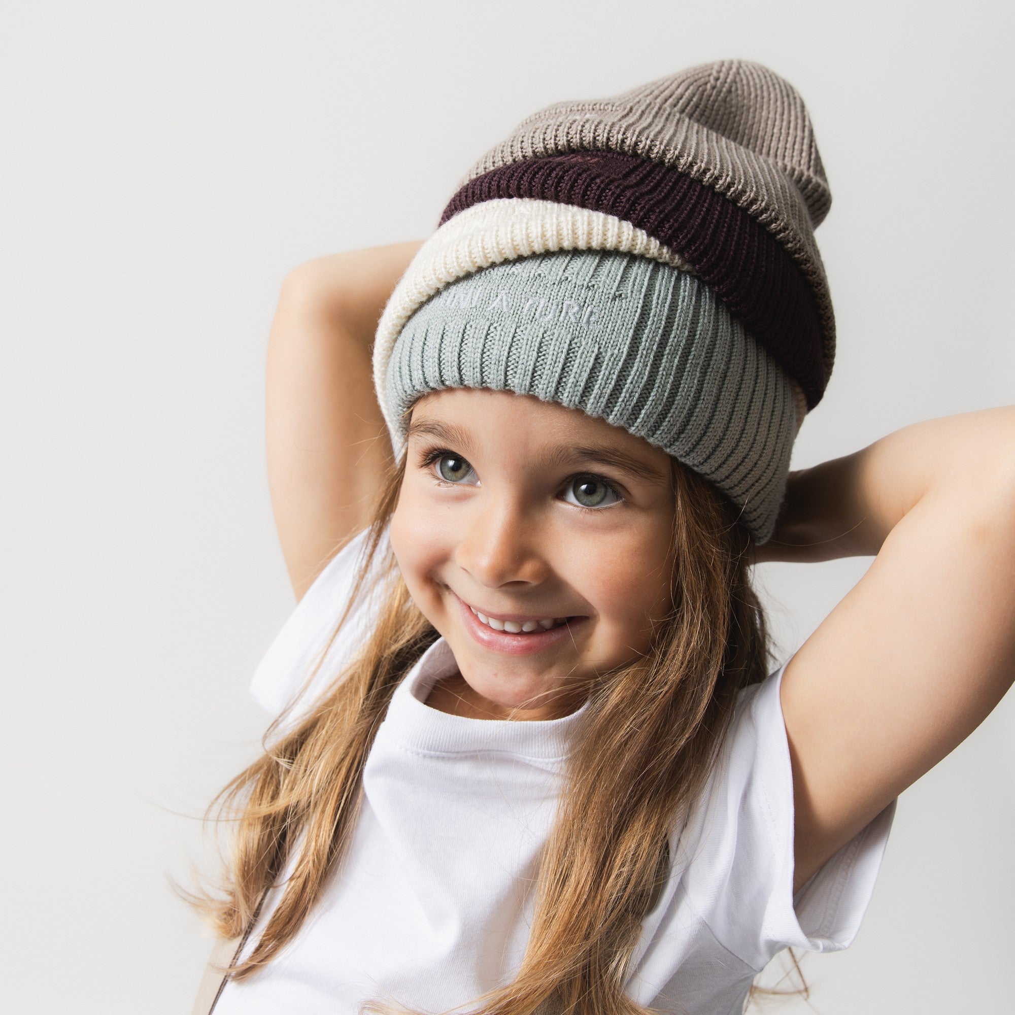 MATBOJE beanie collection stacked on a smiling child's head, showing four colourways in ribbed merino wool with fold-up brims in grey, cream, burgundy and taupe.