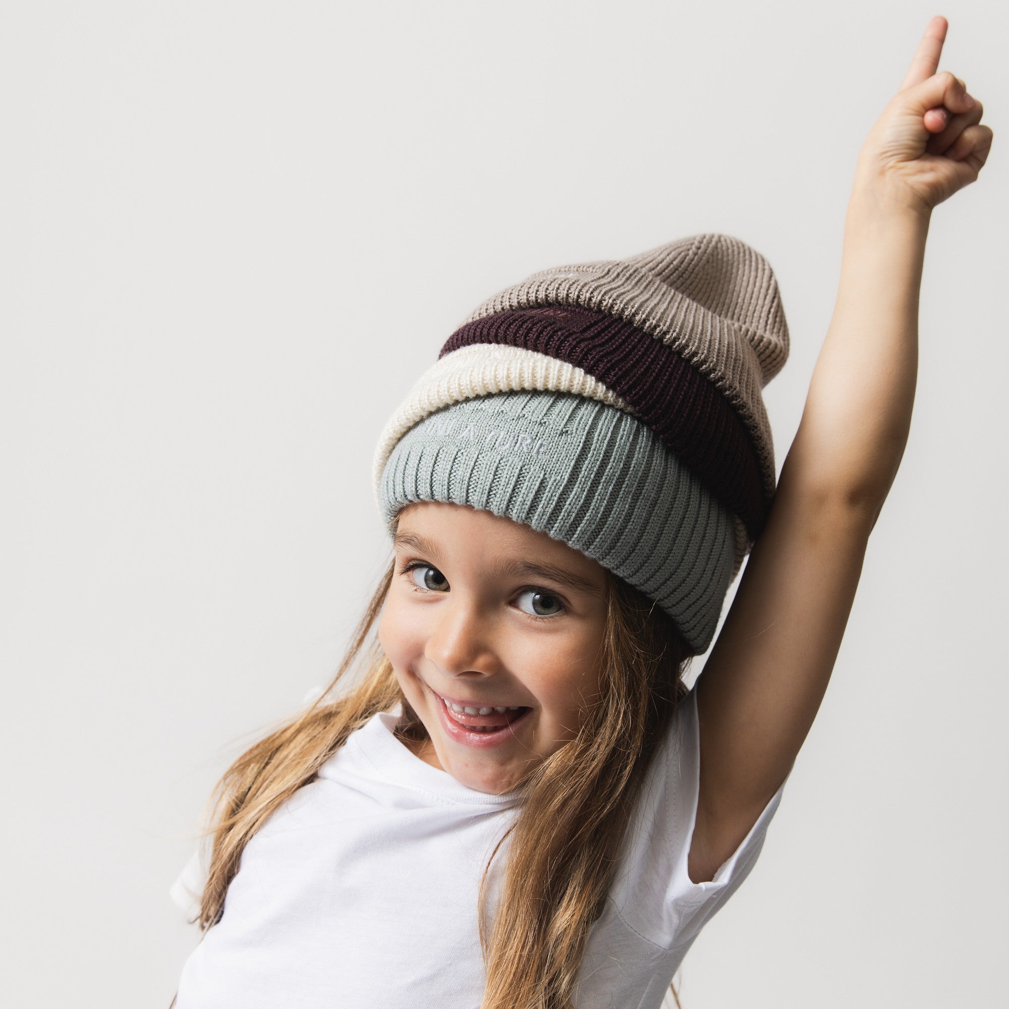 MATBOJE beanie tower stacked on a child's head in four neutral tones, grey-green at the base, cream, plum and taupe, each with a ribbed knit and fold-up brim, worn with a white t-shirt as the child points upward.
