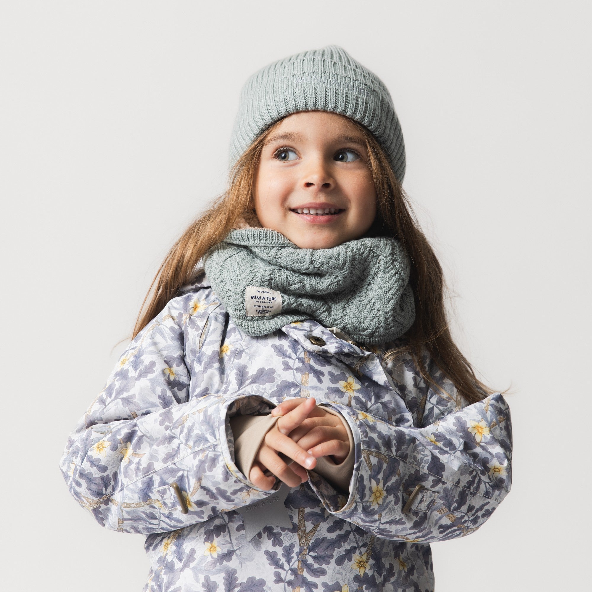 MATBOJE beanie in soft grey-blue merino wool with a ribbed knit, worn with a matching cable-knit neck warmer by a smiling child in a floral jacket.