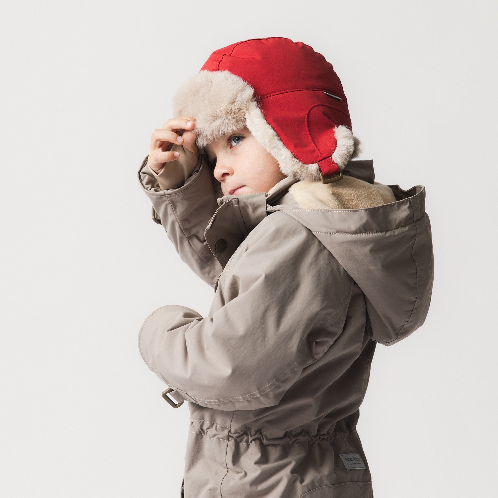 MATCRISTER fleece lined trapper hat in vibrant red with cream faux fur trim, worn by a child adjusting the ear flap while dressed in a taupe winter coat against a neutral studio background.