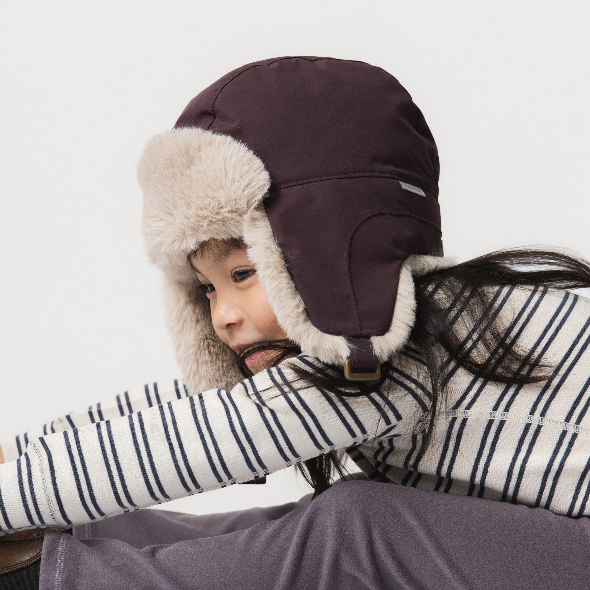 MATCRISTER teddy lined winter hood in dark brown with fluffy fleece ear flaps and chin strap, worn by a child in a striped top.