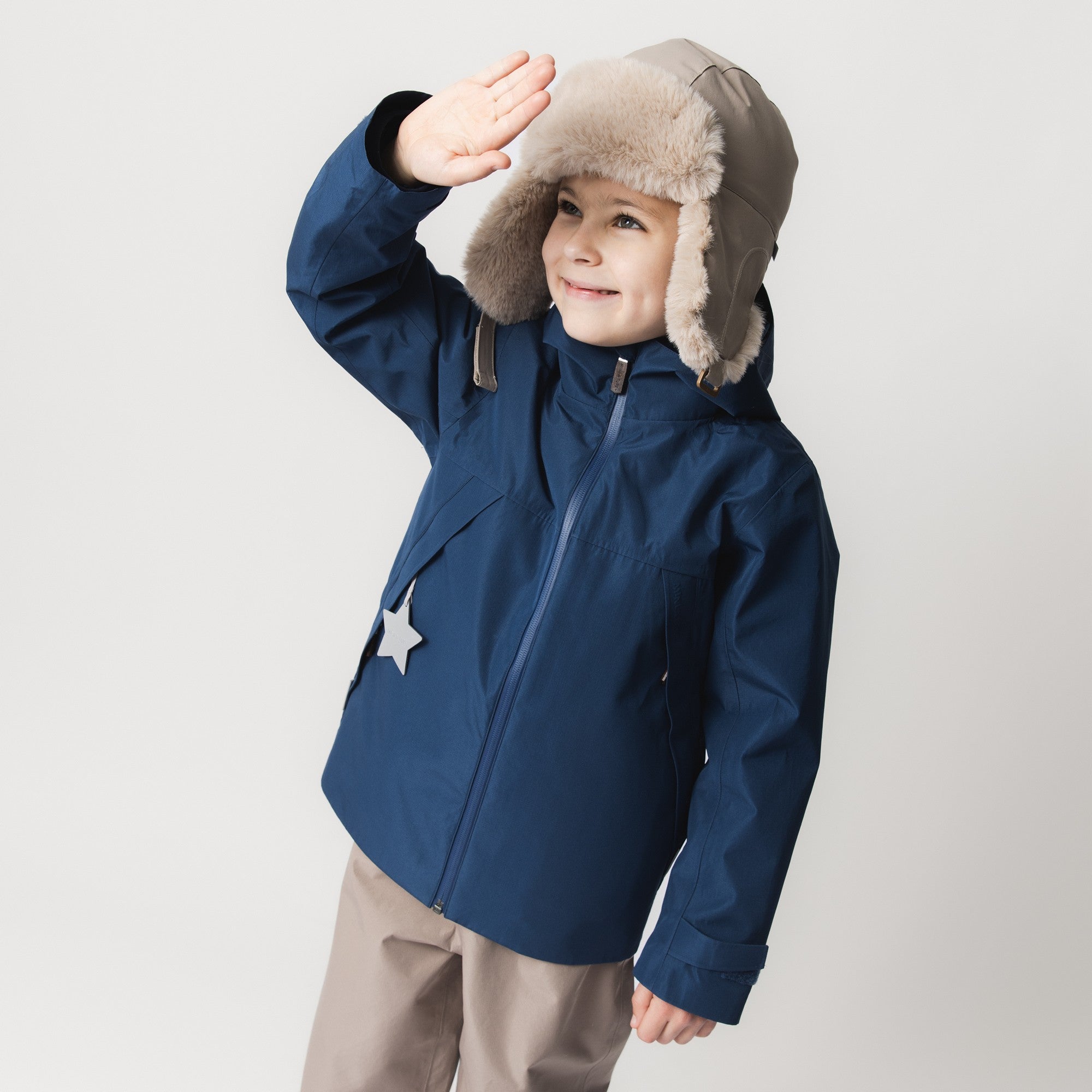 MATCRISTER waterproof trapper hat in stone grey with soft beige fleece lining and ear flaps, worn by a smiling child in a navy blue winter jacket with one hand raised in a wave, photographed against a pale studio background.