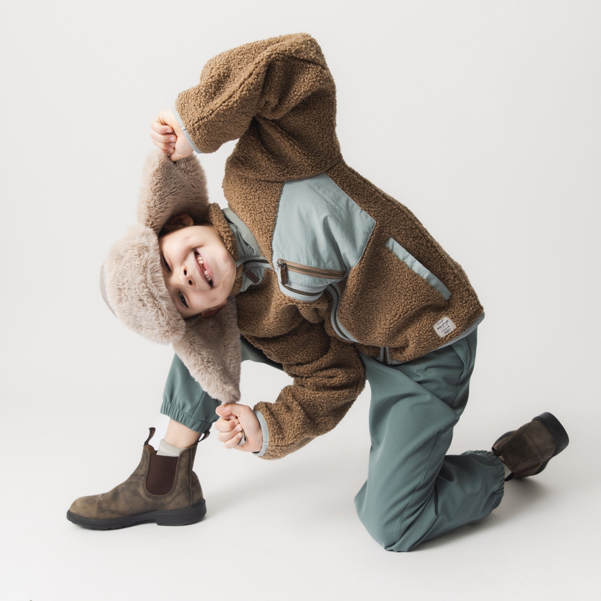 MATCRISTER teddy lined trapper hat in warm brown with matching fleece ear flaps and lining, worn by a smiling child kneeling on the floor in a sage green jacket and trousers against a light studio background.
