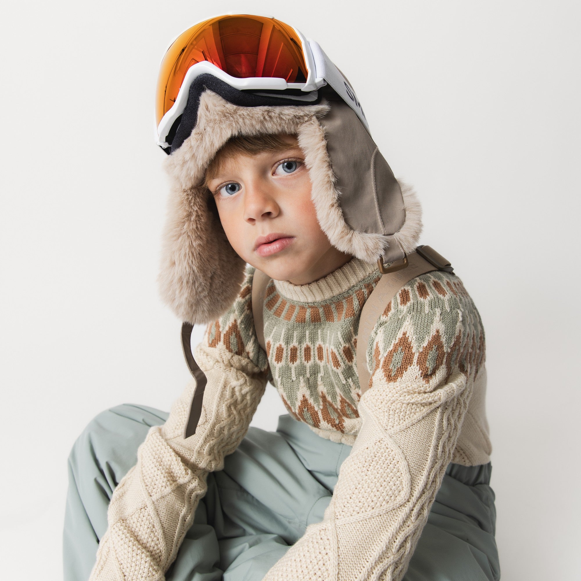 MATCRISTER children's trapper hat in stone grey with plush beige faux fur ear flaps and lining, worn by a child in a patterned jumper and ski goggles, seated against a pale studio background.