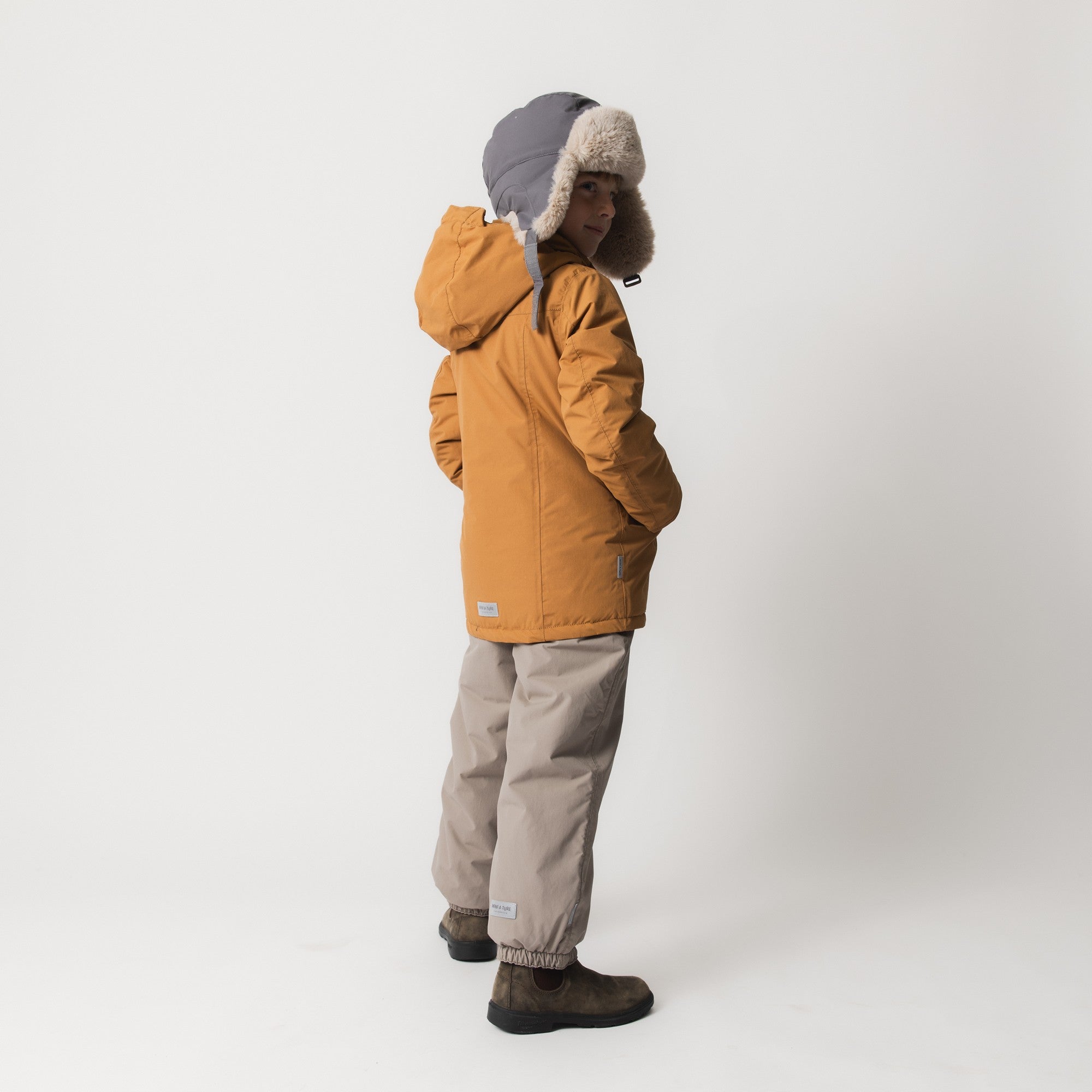 MATCRISTER waterproof trapper hat in slate grey with thick cream fleece lining and ear flaps, worn by a child in a mustard winter jacket and stone trousers, photographed in profile against a pale studio background showing the full-coverage design and shoulder-length protection.