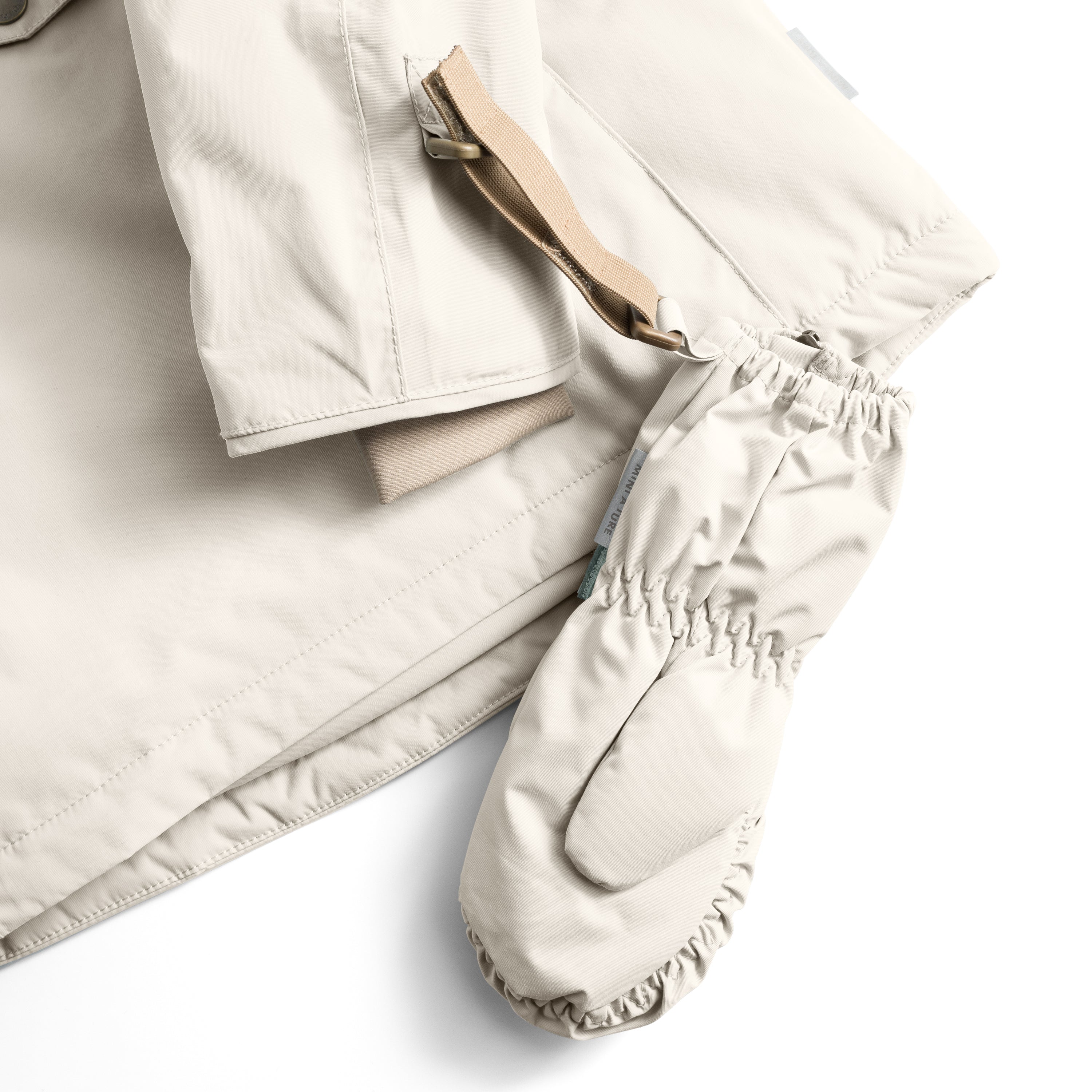 MATCESAR mitten in cream shown threaded onto a matching jacket sleeve via the beige webbing strap and metal D-ring, with the elastic gathered cuff and soft fleece lining visible at the opening, demonstrating how the attachment system keeps mittens secured to outerwear during wear.