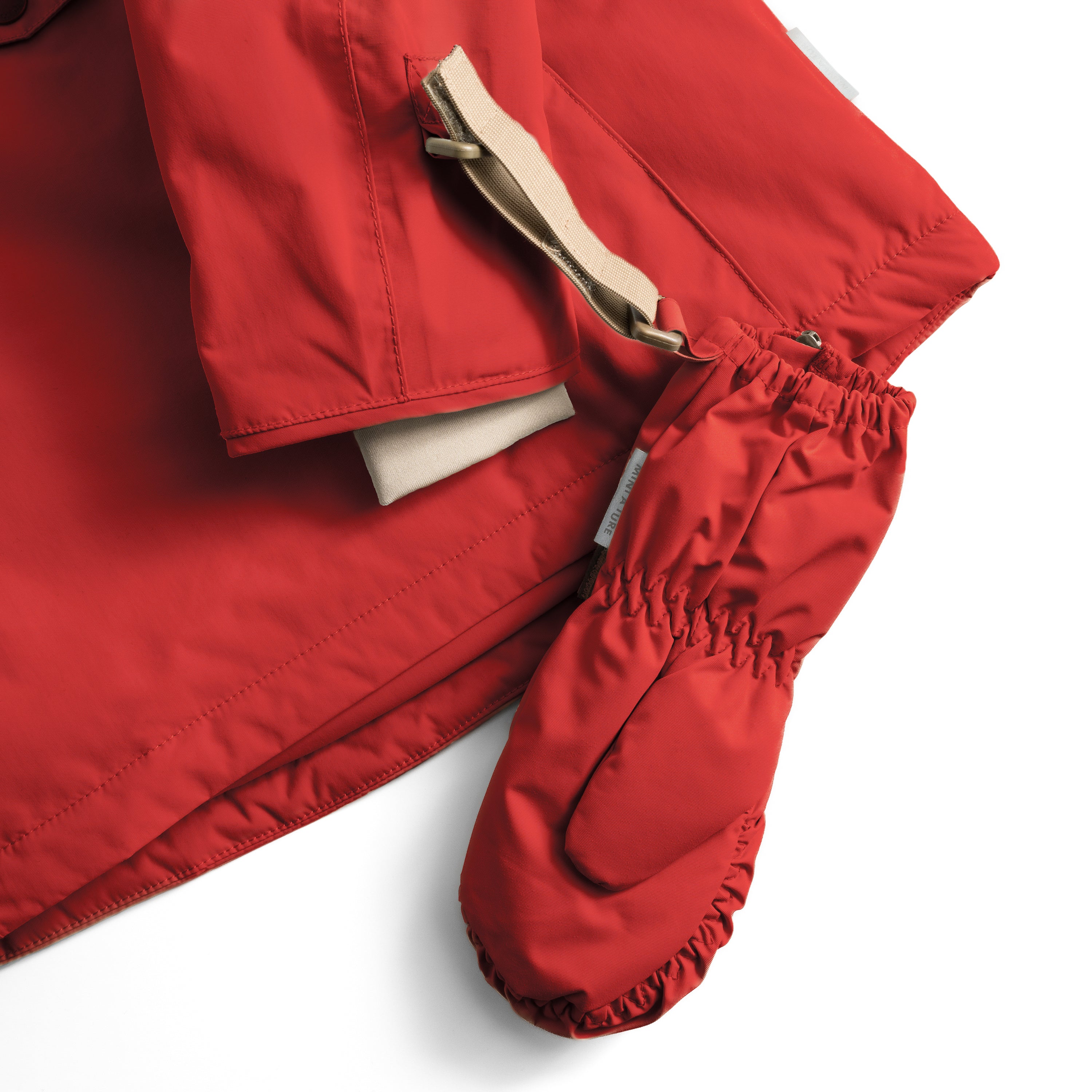MATCESAR mitten in bright red shown in detail with the beige webbing strap threaded through the wrist loop and attached to a matching red jacket sleeve, highlighting the D-ring attachment system and elastic gathered cuff with fleece lining visible at the opening.