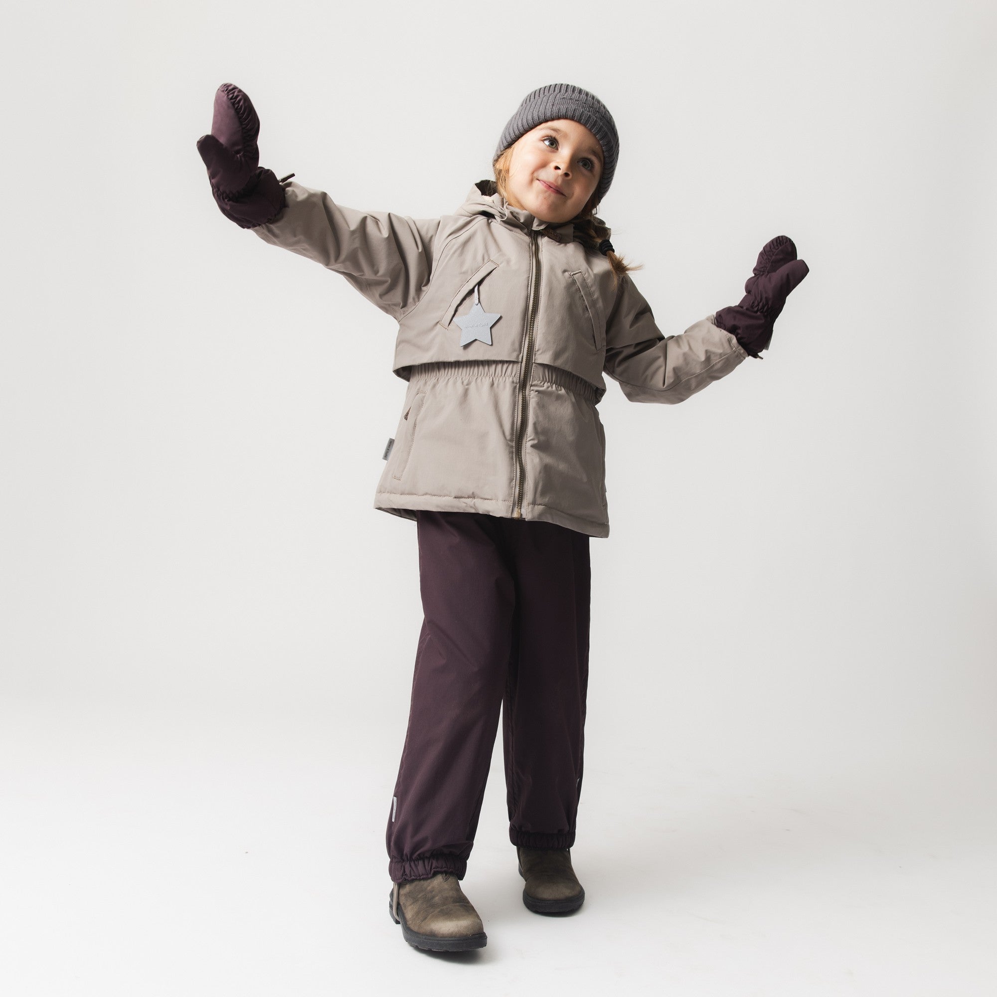 Child wearing MATCESAR waterproof mittens in plum with a beige winter jacket, grey knitted beanie and dark trousers, arms raised in a playful pose against a white studio background.