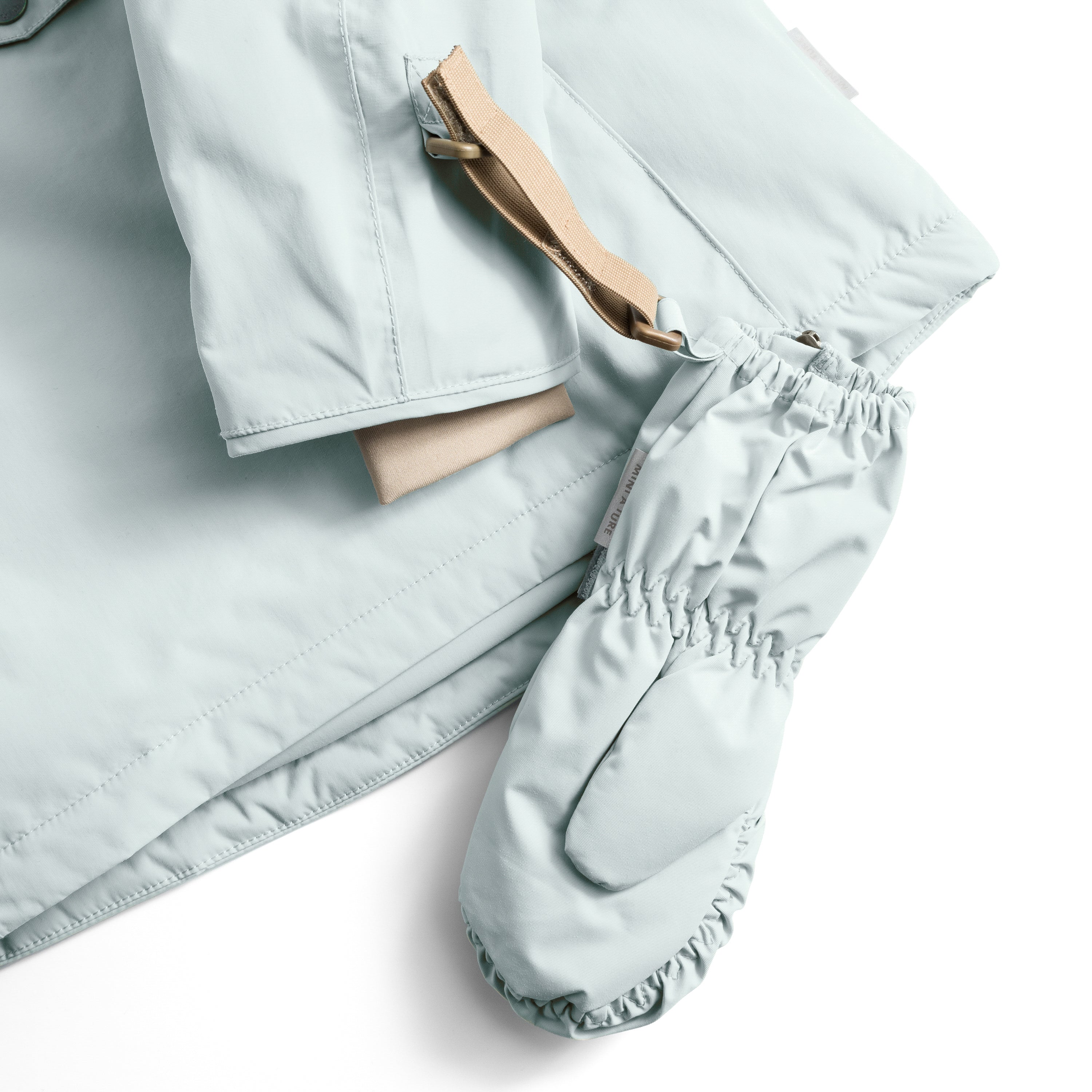 MATCESAR mitten in pale blue shown threaded onto a matching jacket sleeve via the beige webbing strap and D-ring, demonstrating the attachment system that prevents mittens from being lost, with the elastic gathered cuff and soft fleece lining visible at the opening.