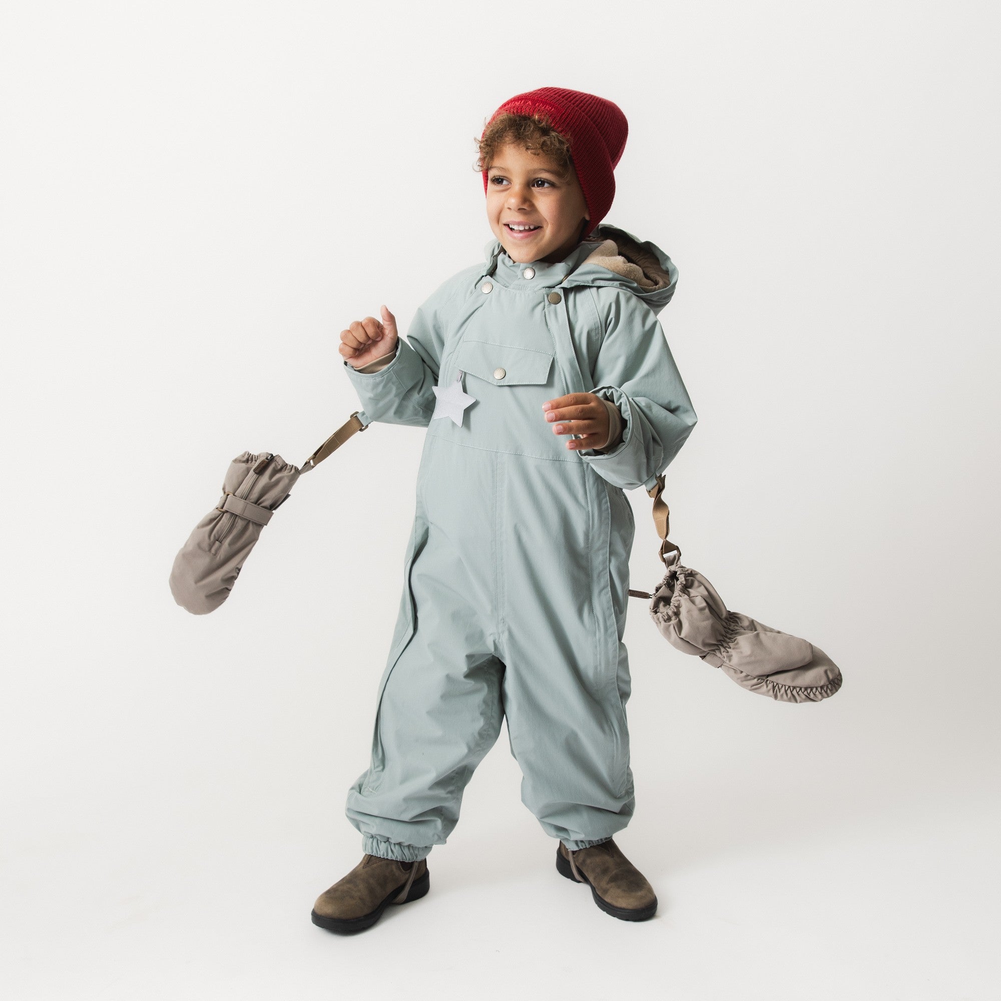 Child wearing MATCESAR waterproof mittens in grey taupe attached by beige wrist straps to a pale blue snowsuit, paired with a burgundy knitted beanie, holding one mitten by the strap in a playful gesture against a white studio background.