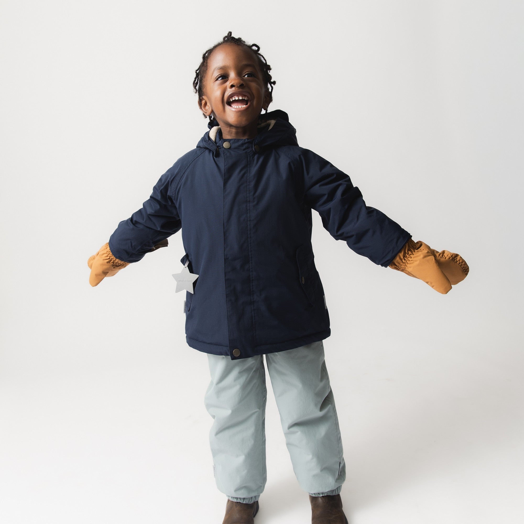MATCORDT fleece lined mittens in caramel brown worn by a young child in a navy jacket, with arms outstretched in joyful movement, showing the secure fit at the wrist and the freedom of grip the THINSULATE padded construction provides during active winter play.
